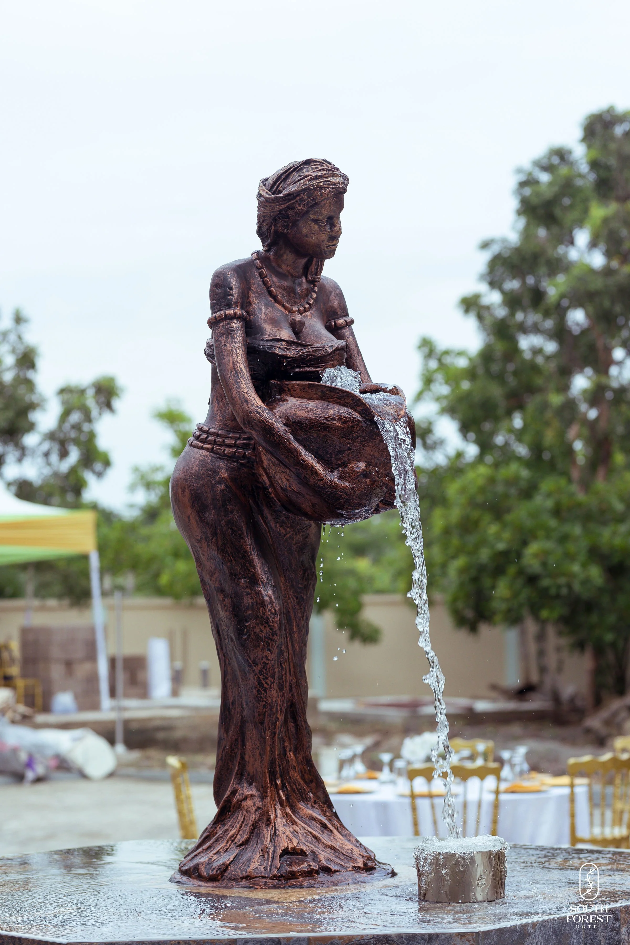 A bronze statue of a woman pouring water from a vessel into a fountain, surrounded by trees and outdoor setup.