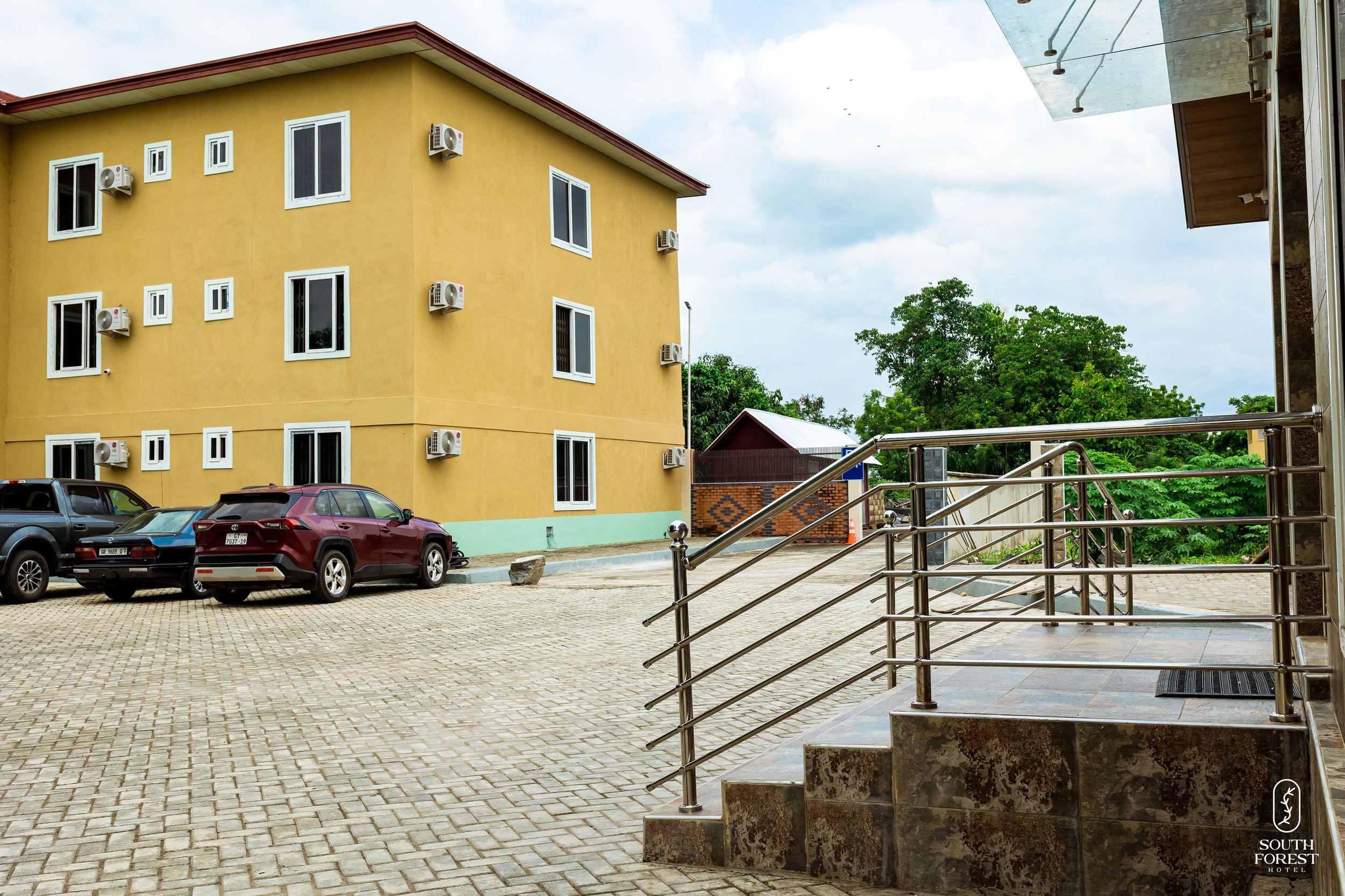 Parking lot outside a yellow hotel building with several windows and air conditioning units, stairs with metal railing leading to the entrance, a few parked cars, some greenery and trees in the background, and cloudy sky.