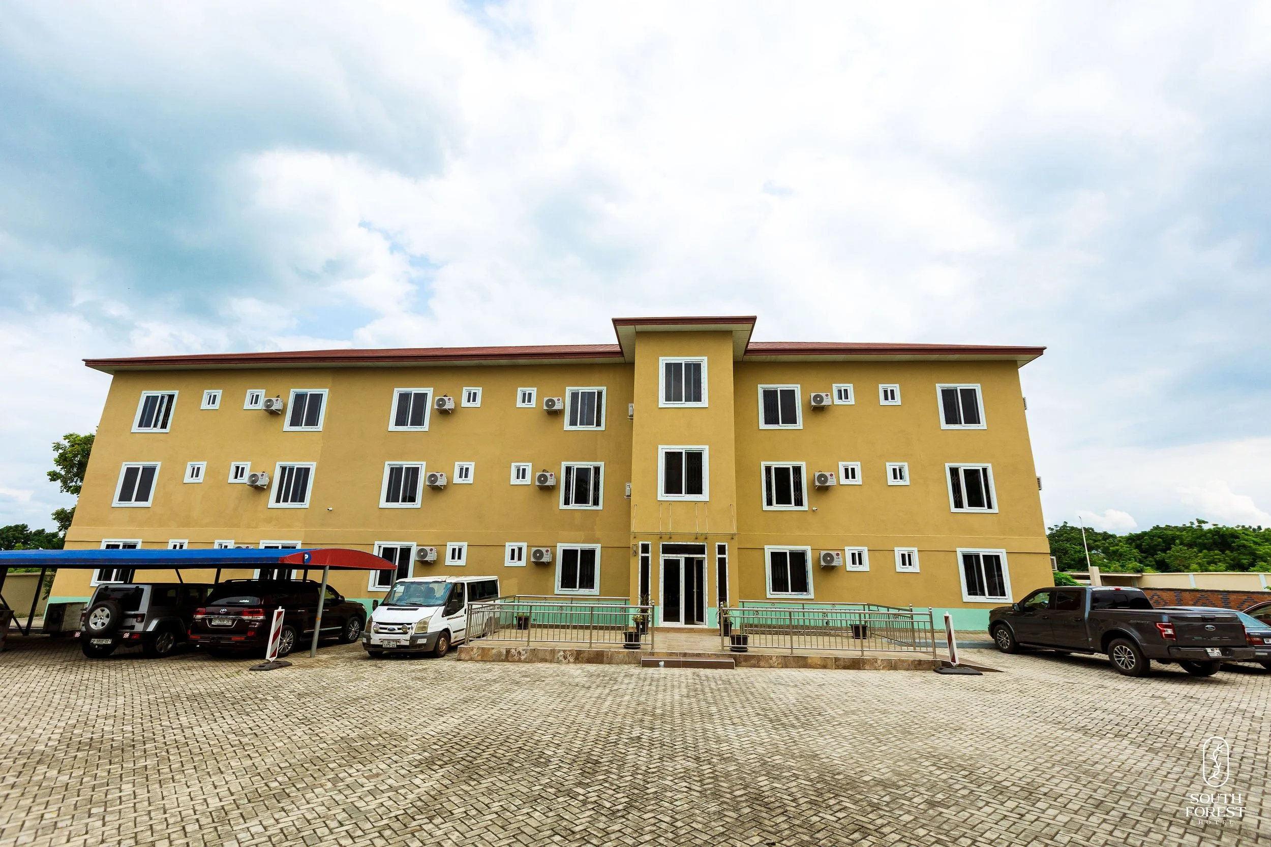 A yellow three-story building with multiple windows and air conditioning units, surrounded by a parking lot with several vehicles parked underneath a carport, under a partly cloudy sky.