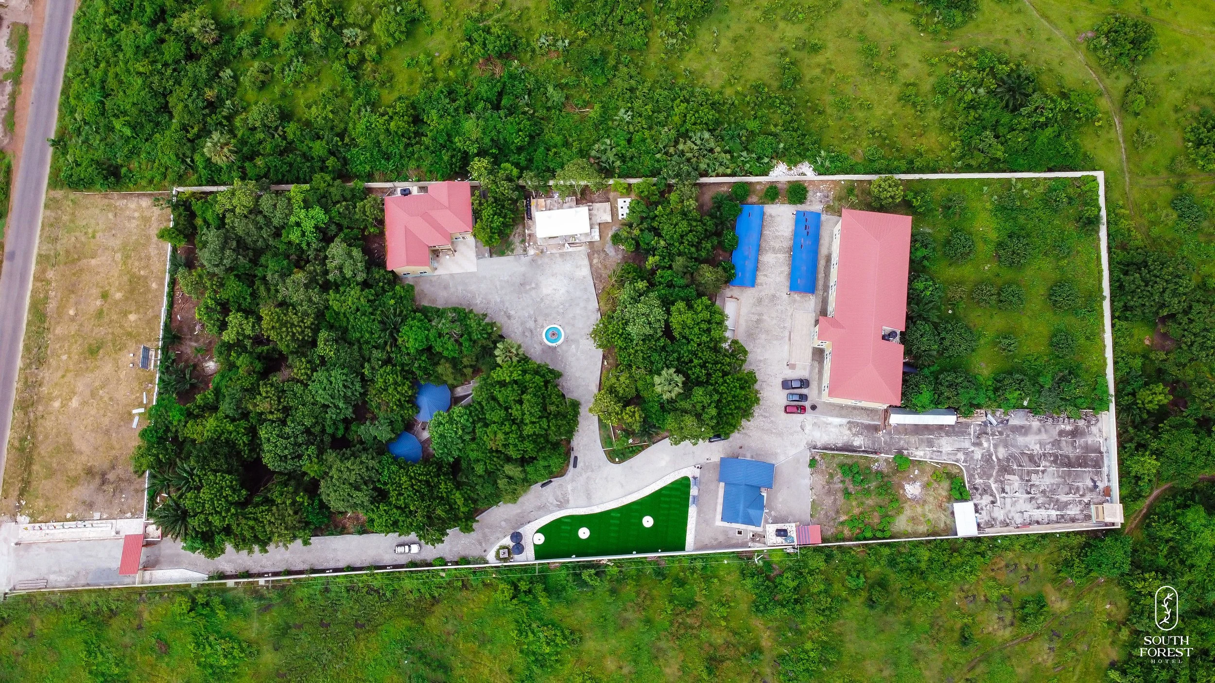 Aerial view of a property with multiple buildings, greenery, and a parking area, surrounded by trees and open land.