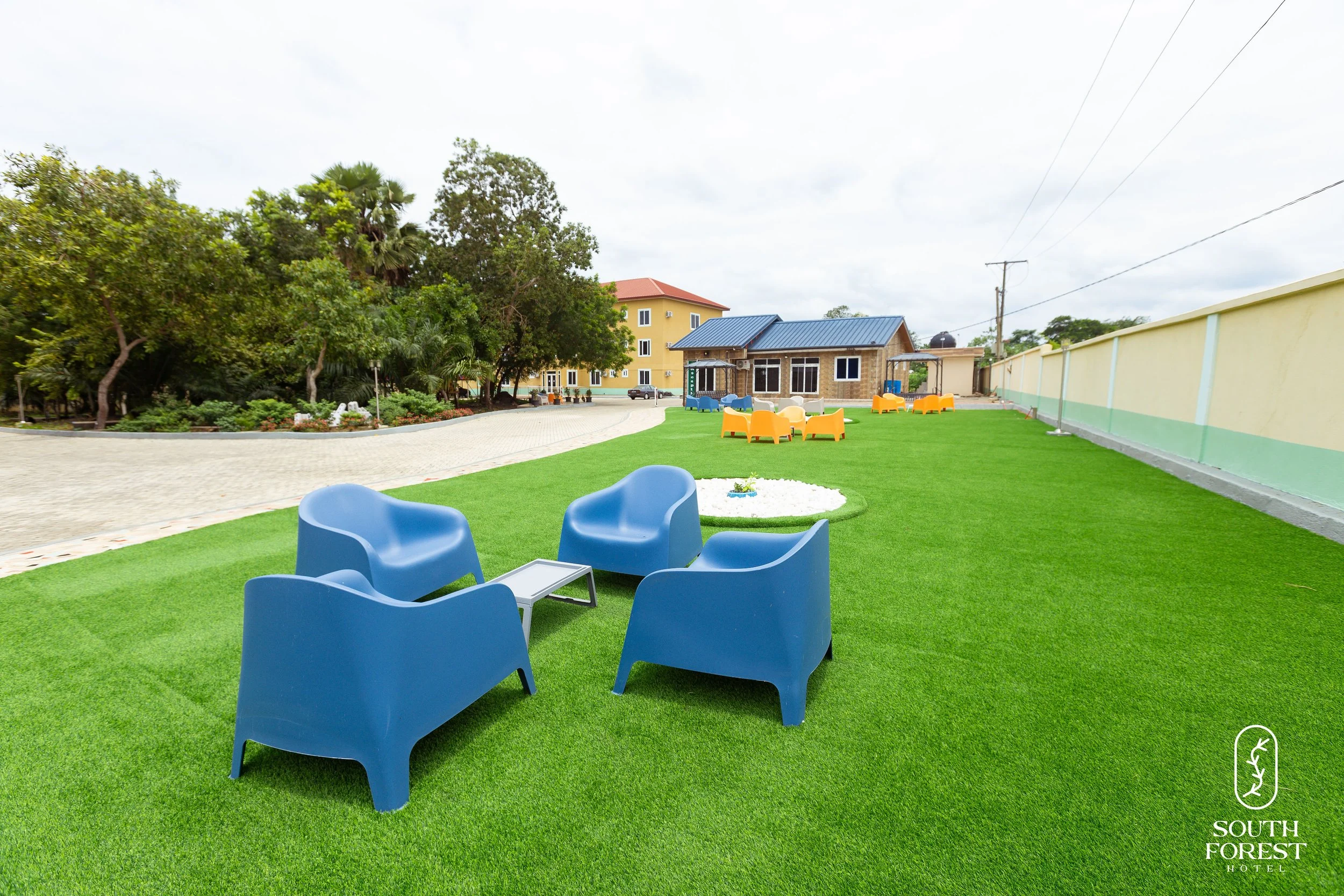 Outdoor seating area with blue chairs and a small table on a well-maintained lawn. In the background, there are colorful seating arrangements, trees, and buildings, including a hotel with a logo in the bottom right corner that reads "South Forest Hot
