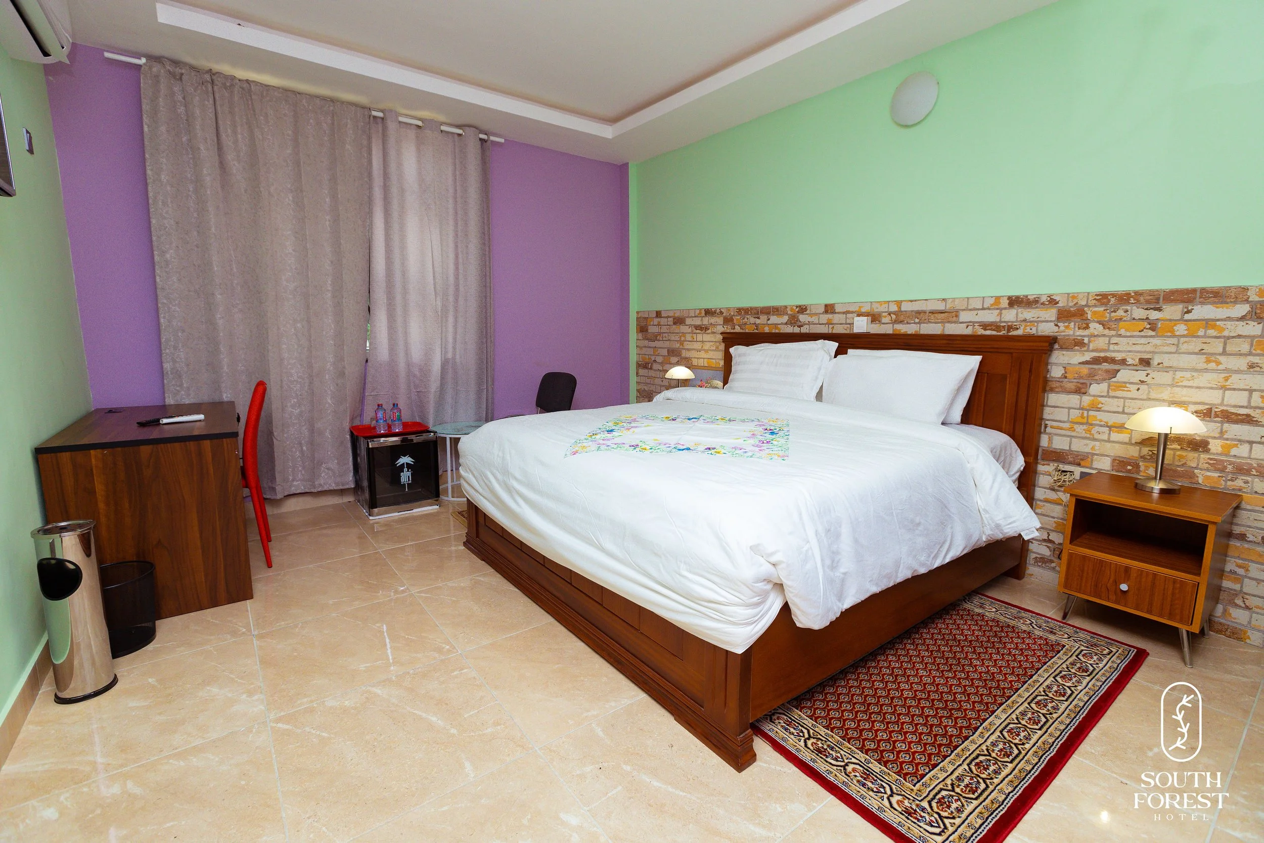 Hotel room with a large bed, nightstand with lamp, wooden desk, and curtains over the window, decorated with a brick accent wall and a small rug on tan tile floor.