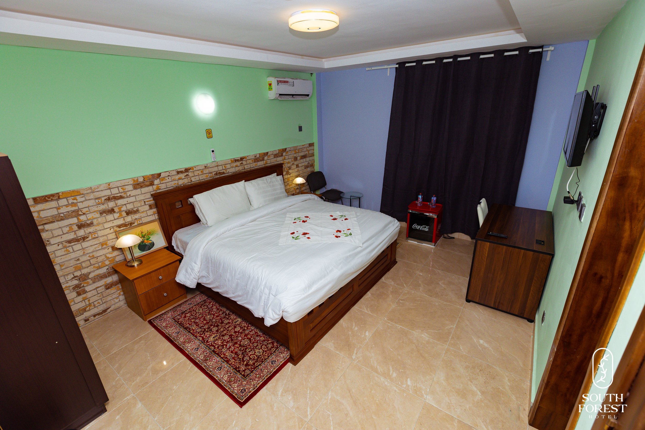 Hotel room with a large bed, wooden headboard, nightstands, lamps, a small rug, a side table, a flat-screen TV, and curtains covering a window. The room has green and purple walls, a brick accent wall, and beige tiled flooring.