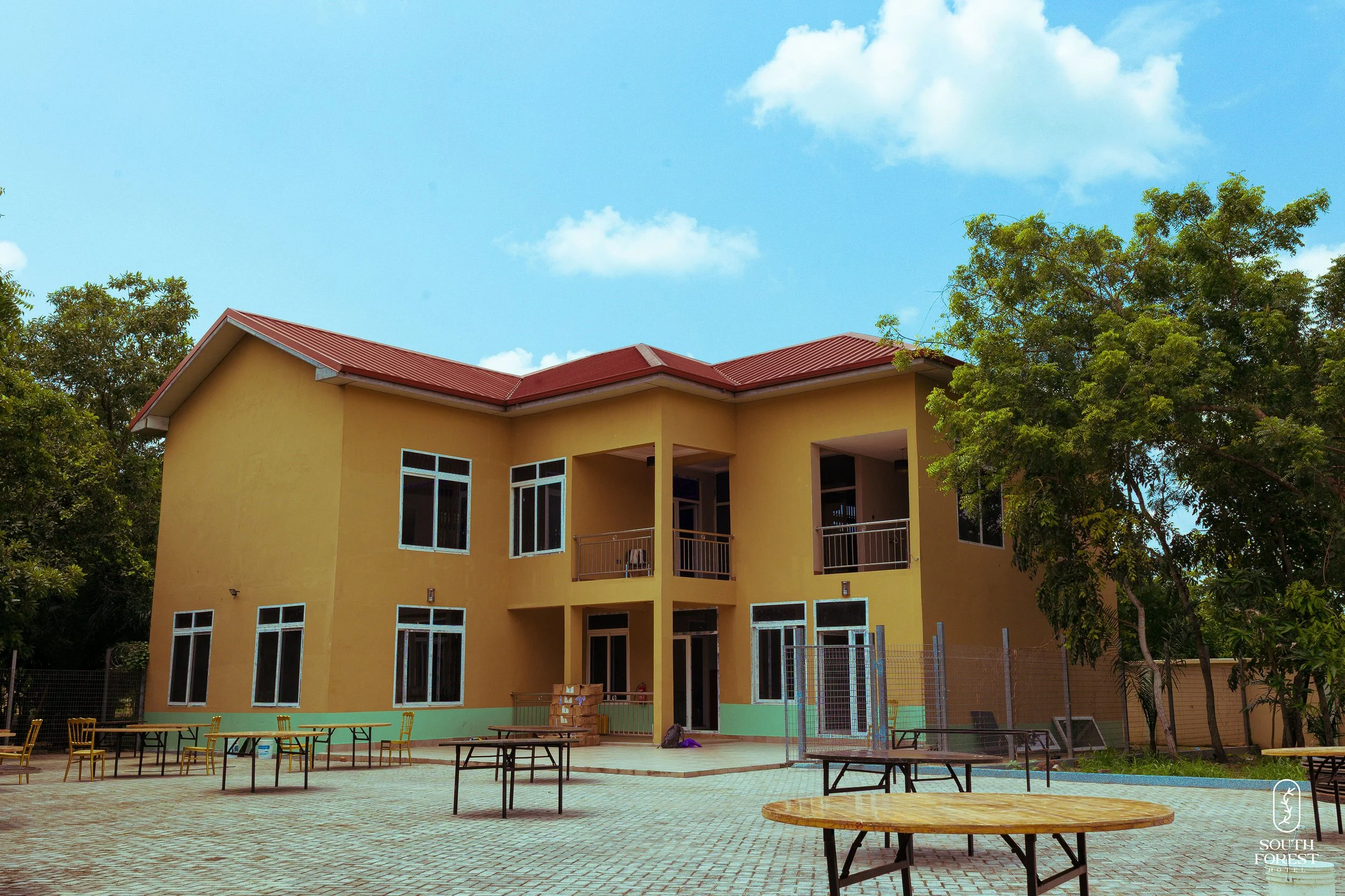 A two-story yellow building with a red roof, surrounded by trees, under a blue sky with white clouds, with outdoor tables and chairs in the foreground.