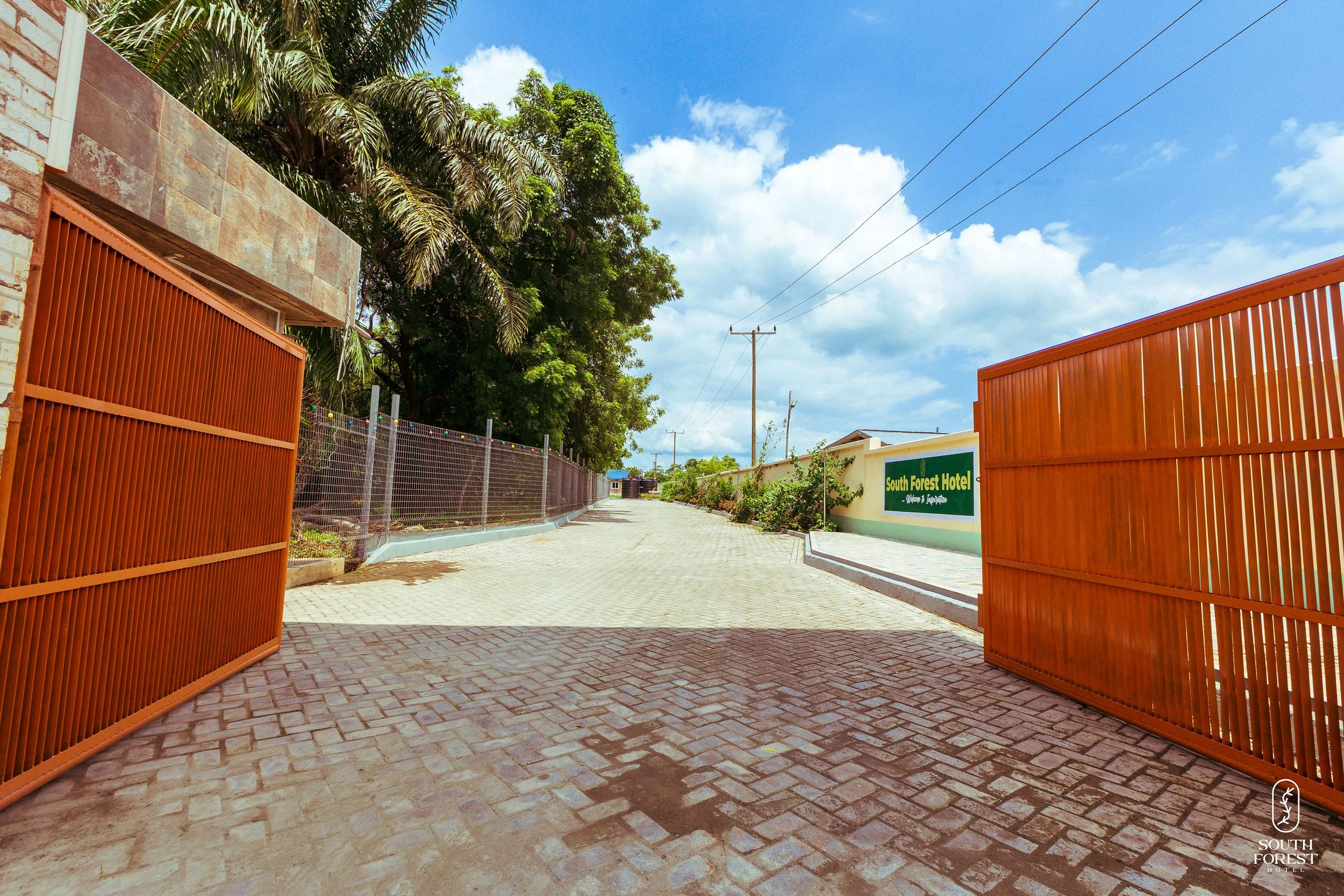 Entrance gates to South Forest Hotel with open rust-colored gates, paved driveway, green signage, and tropical trees under a partly cloudy sky.