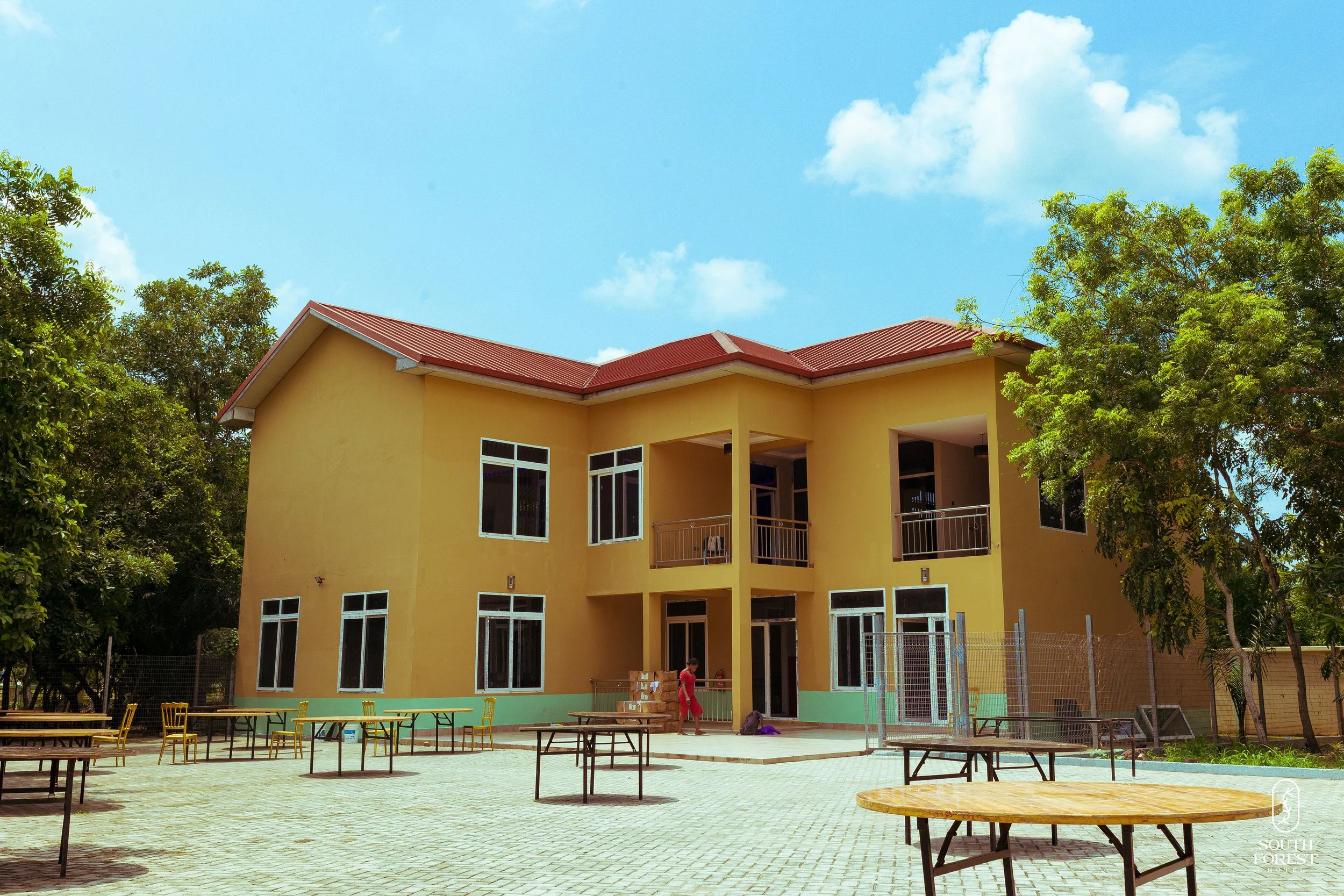 A yellow two-story building with red roof, surrounded by trees, and an outdoor area with tables and chairs under a bright blue sky.