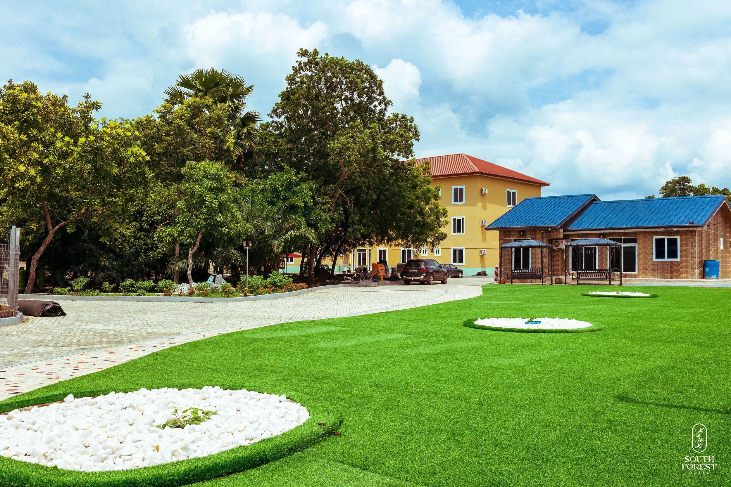 A landscaped outdoor area with green grass, trees, and plants. There are two residential buildings, one yellow with a red roof and the other brick with a blue roof. The area includes a paved walkway and some parked cars, with a blue sky and clouds ov