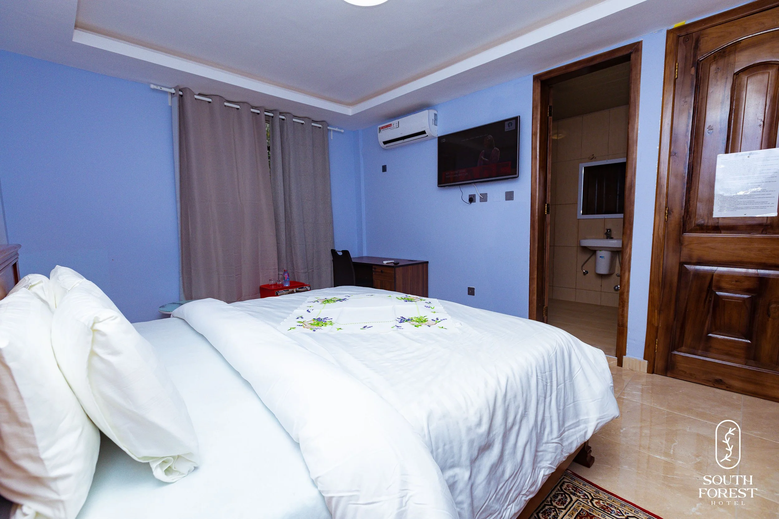 Hotel room with a bed, beige curtains, a small desk, a wall-mounted TV, and an entrance to a bathroom with a sink. The room has light blue walls and wooden doors.