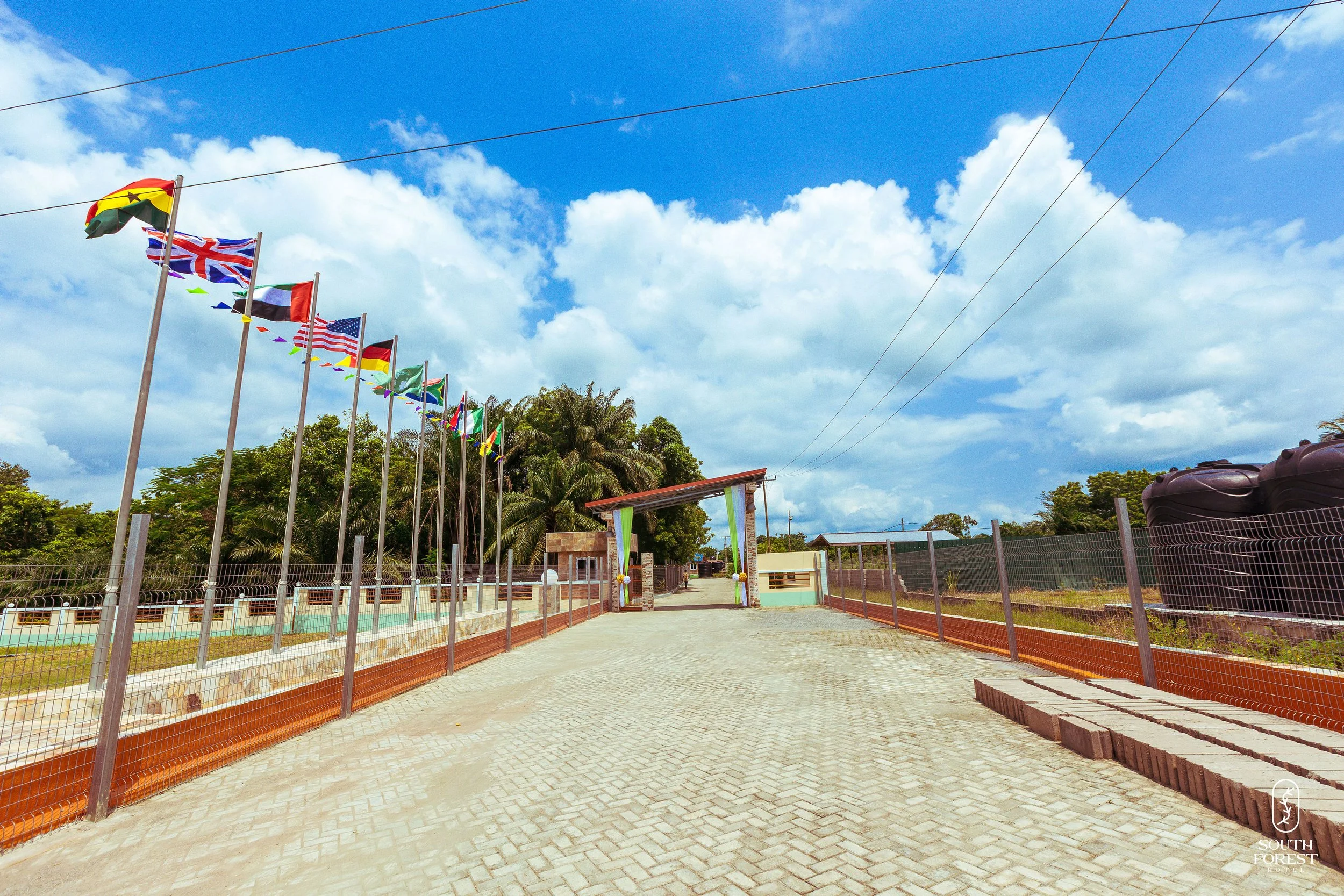 A paved pathway at an outdoor location with multiple flags from different countries on poles on the left, a fence on both sides, trees in the background, a small building or gate structure ahead, and blue sky with scattered clouds overhead.