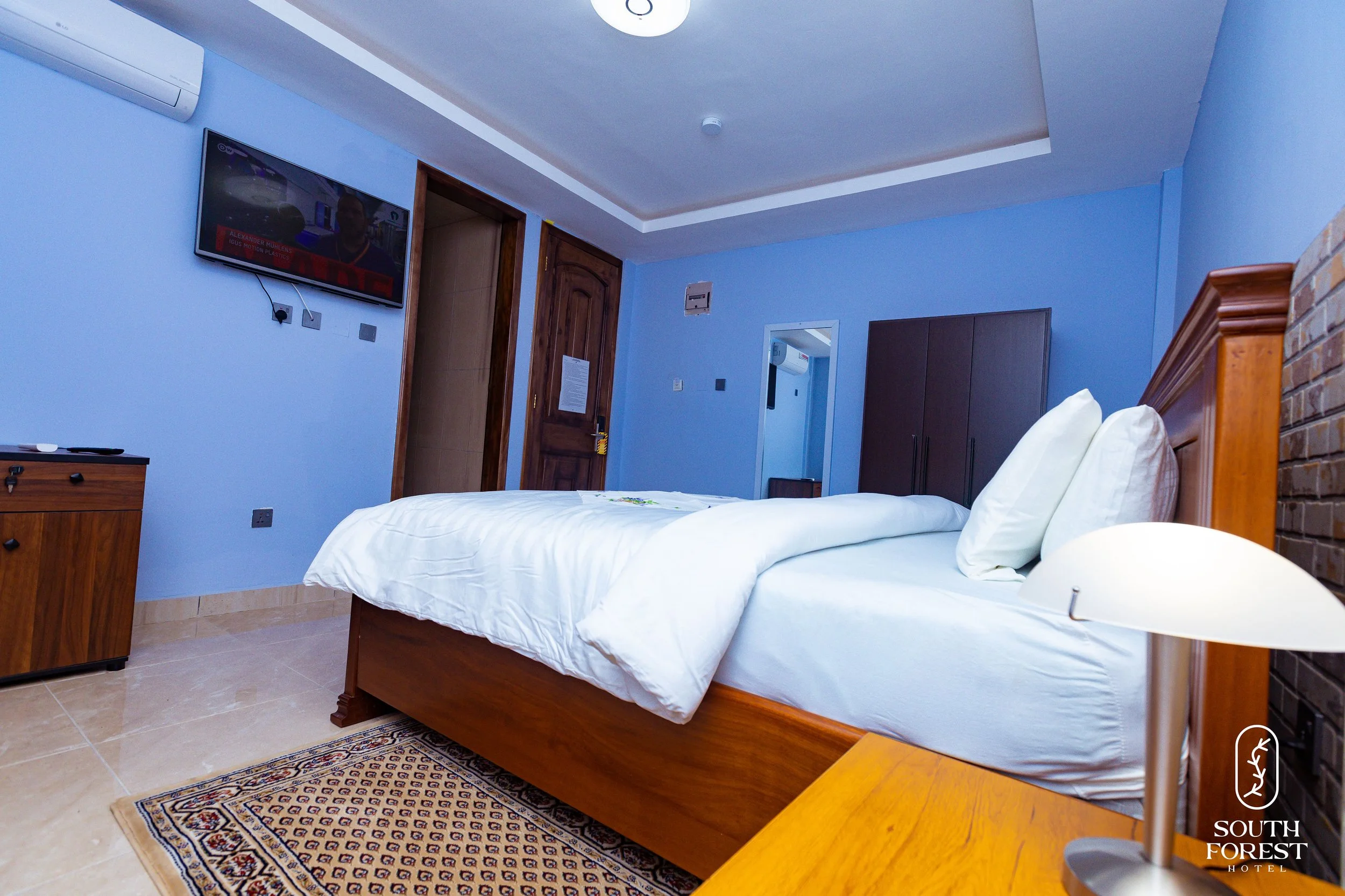 A hotel room with a double bed, white bedding and pillows, a nightstand with a white lamp, a wooden wardrobe, a small cabinet, a wall-mounted TV, blue painted walls, a wooden door, and a religious symbol on the wall at the South Forest Hotel.
