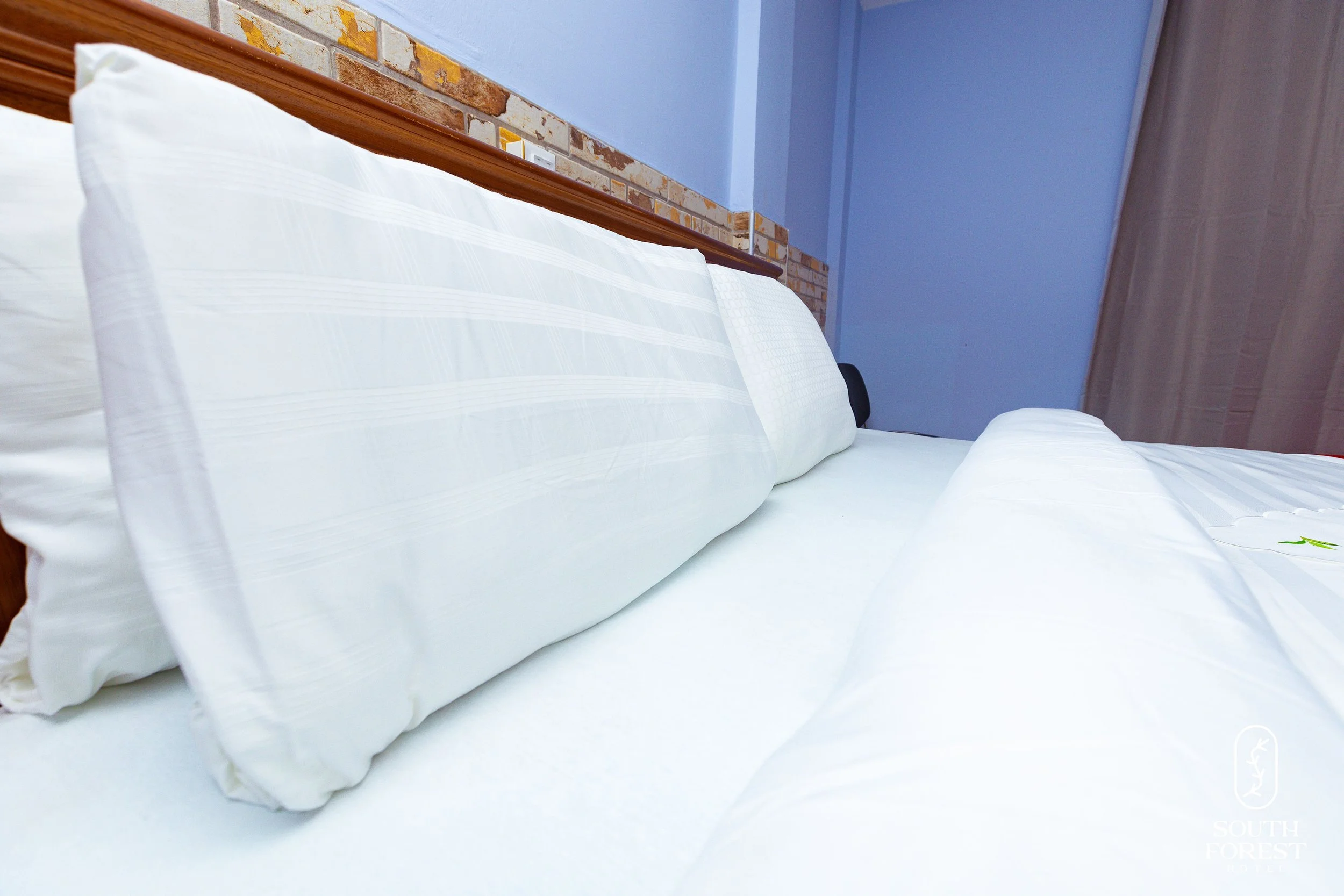White pillows and bed sheets on a neatly made bed with a wooden headboard, a brick accent wall above, and a beige curtain in the background.