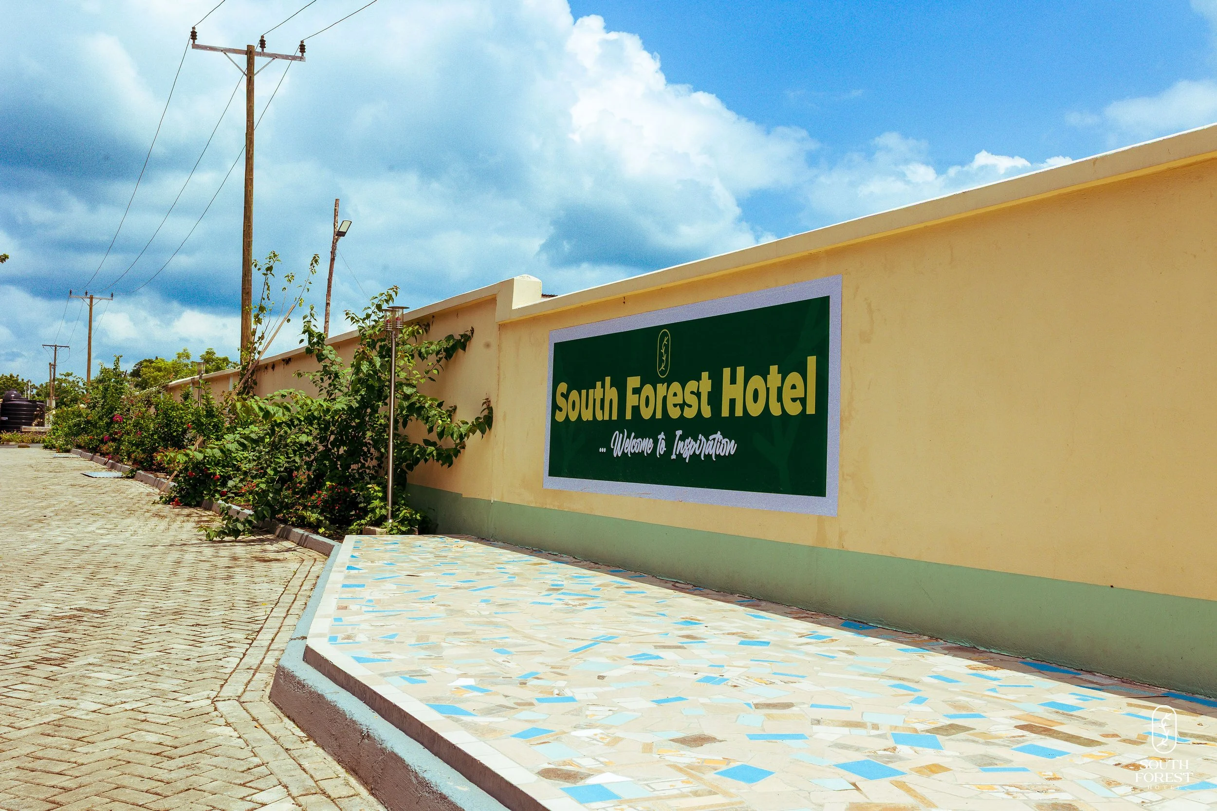 Exterior of South Forest Hotel with a large green sign, yellow wall, concrete sidewalk, and cloudy blue sky.