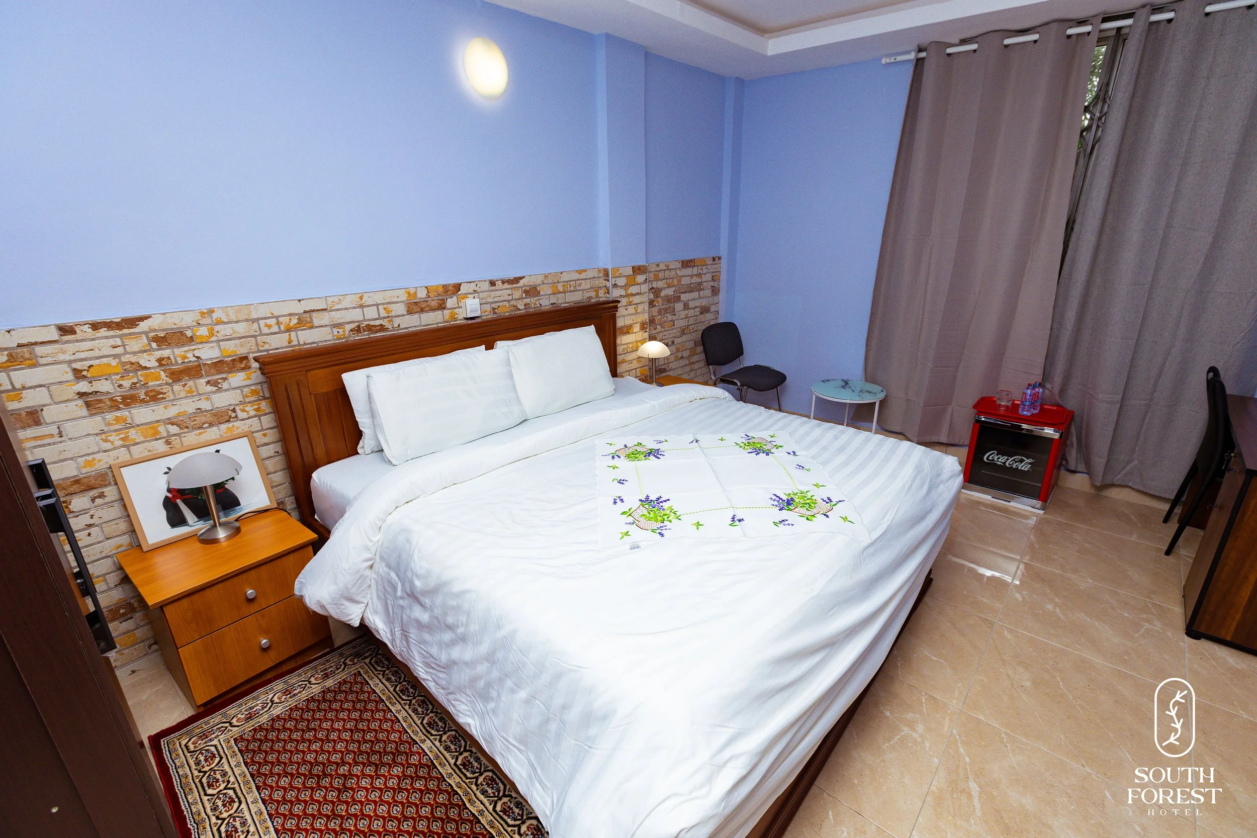 Hotel room with a large bed, brick accent wall, nightstand with a lamp, chairs, side table, curtains, and a mini fridge.