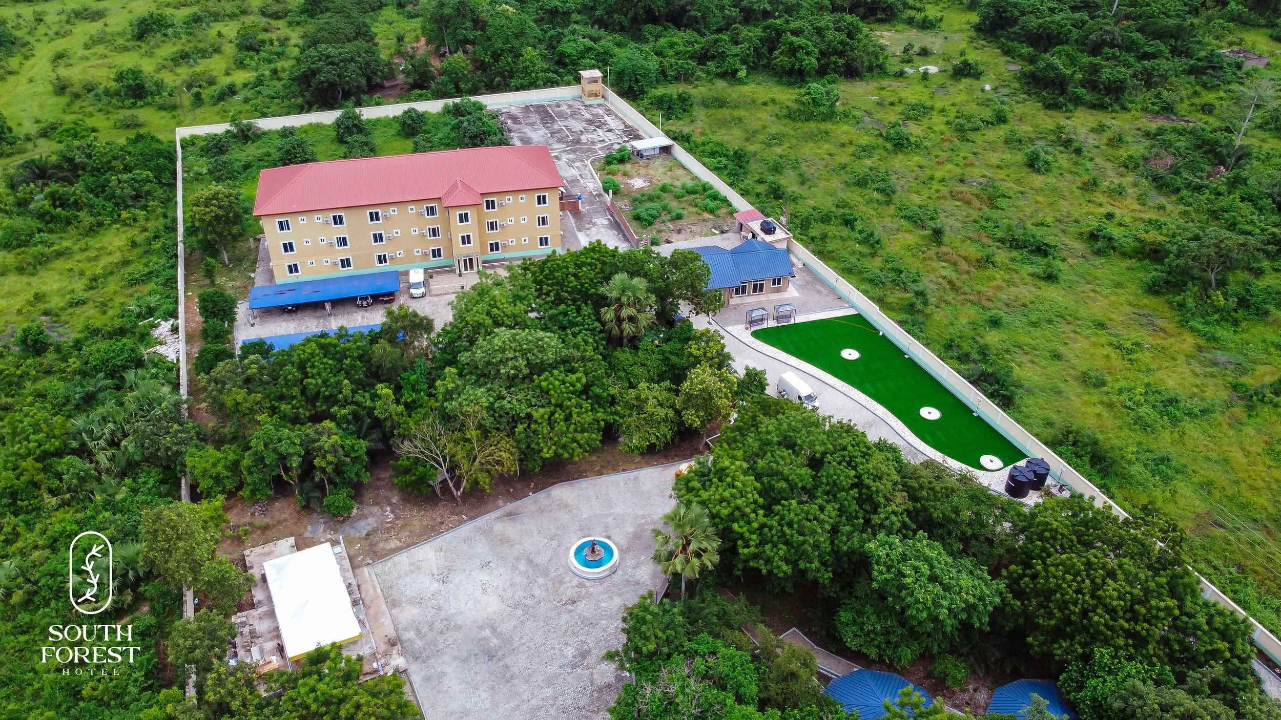 Aerial view of a large beige hotel with a red roof, surrounded by green trees, with a small blue-roofed building and a fenced sports area with three white circular sections and black water tanks nearby. There is a circular fountain and a white tent a