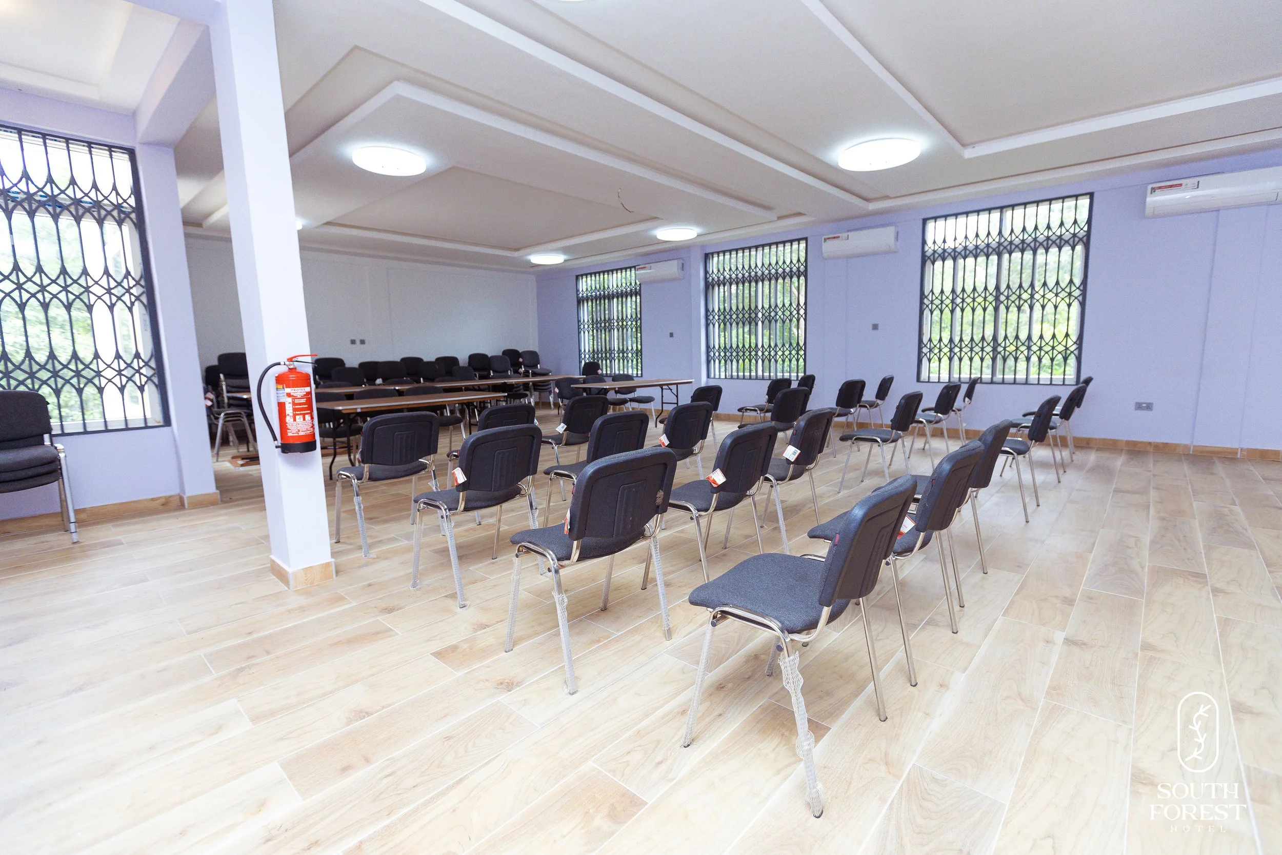 Empty conference room with black chairs arranged in rows, large windows with security bars, wooden floor, air conditioning units, and a fire extinguisher on a white pillar.