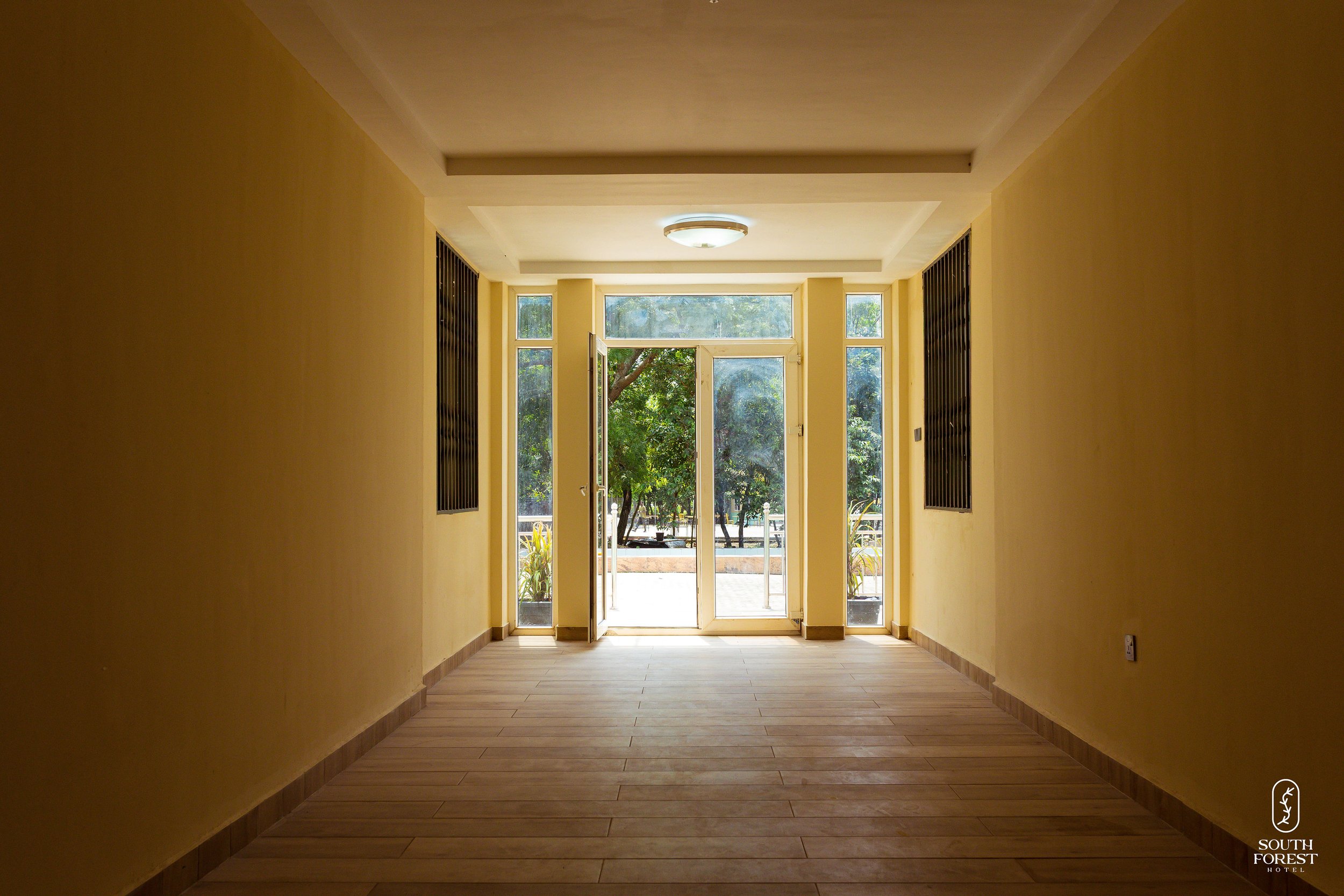 Empty hallway with large glass doors leading outside, sunlight streaming in, yellow walls, wooden flooring, South Forest Hotel logo in bottom right corner.