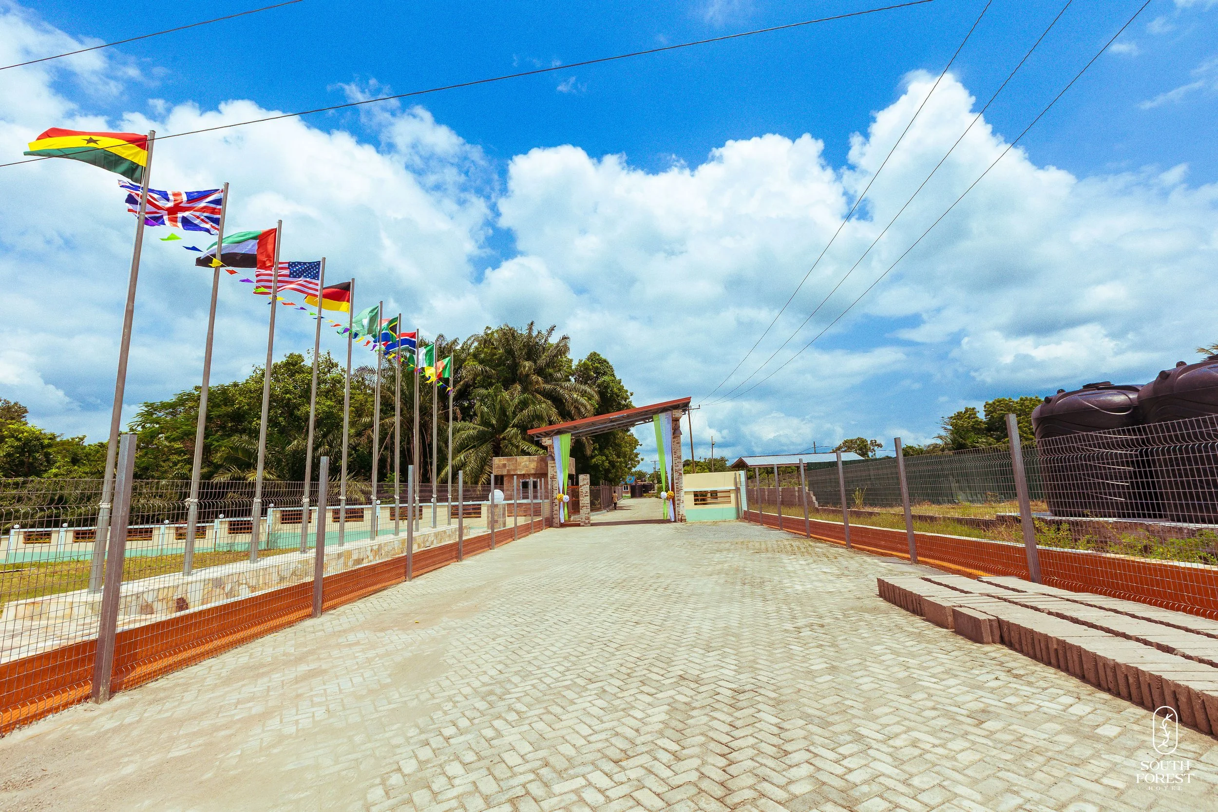 A paved walkway leading to a gated entrance decorated with flags of various countries on poles. The sky is partly cloudy with blue sky visible. Green trees and palm trees are in the background.