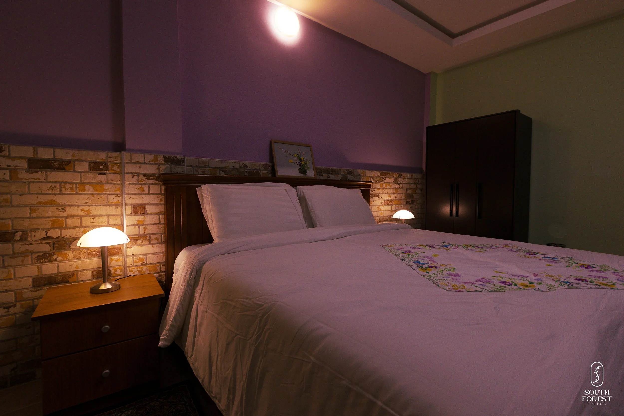 A bedroom with a bed featuring white bedding and pillows, a wooden headboard, two bedside lamps, a framed picture of flowers on the wall, a black wardrobe, and a brick accent wall, with purple and mint green painted sections on the walls.