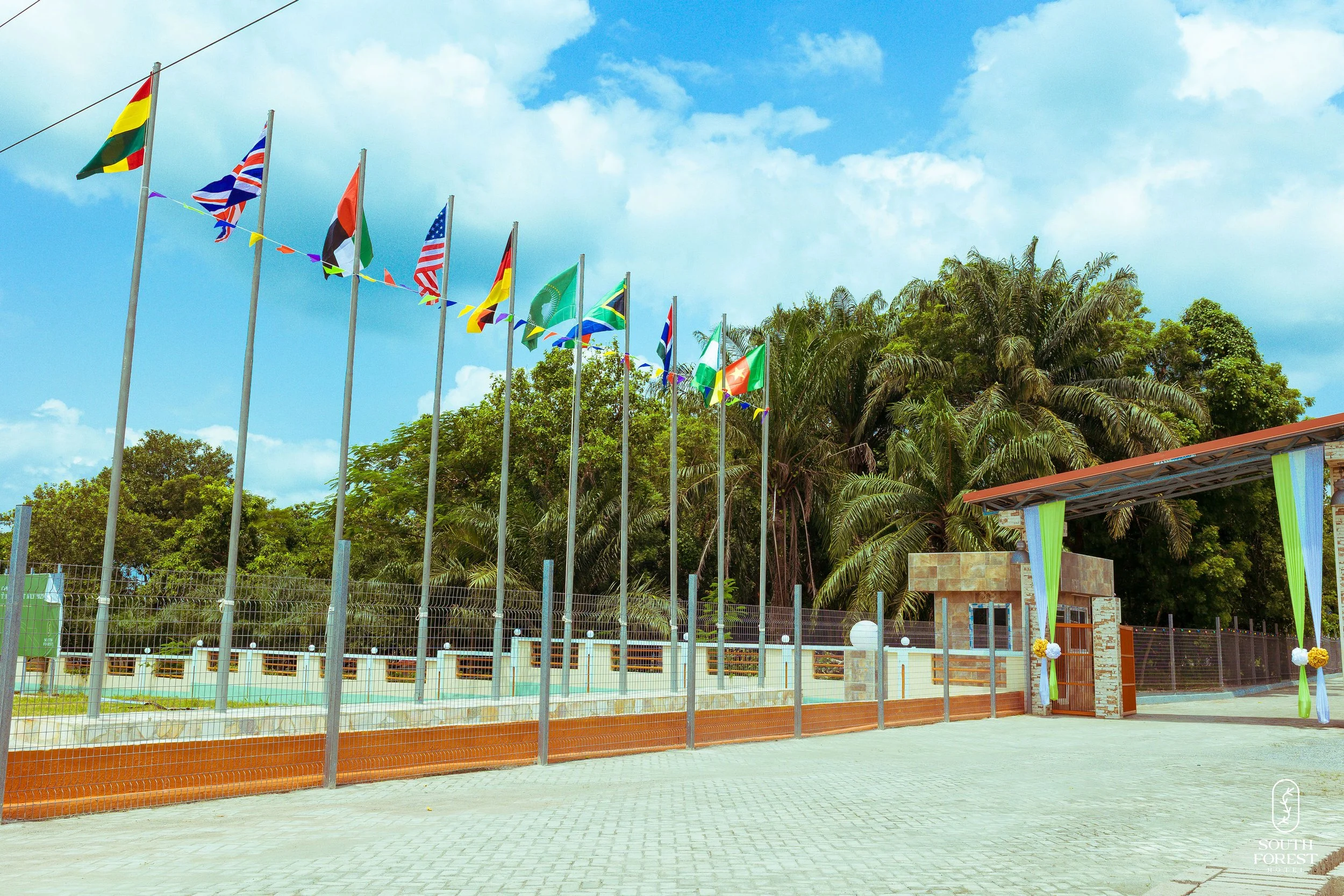 Flag poles with various national flags, a gated entrance decorated with green drapes and flower arrangements, palm trees and other greenery in the background, and a partly cloudy sky.