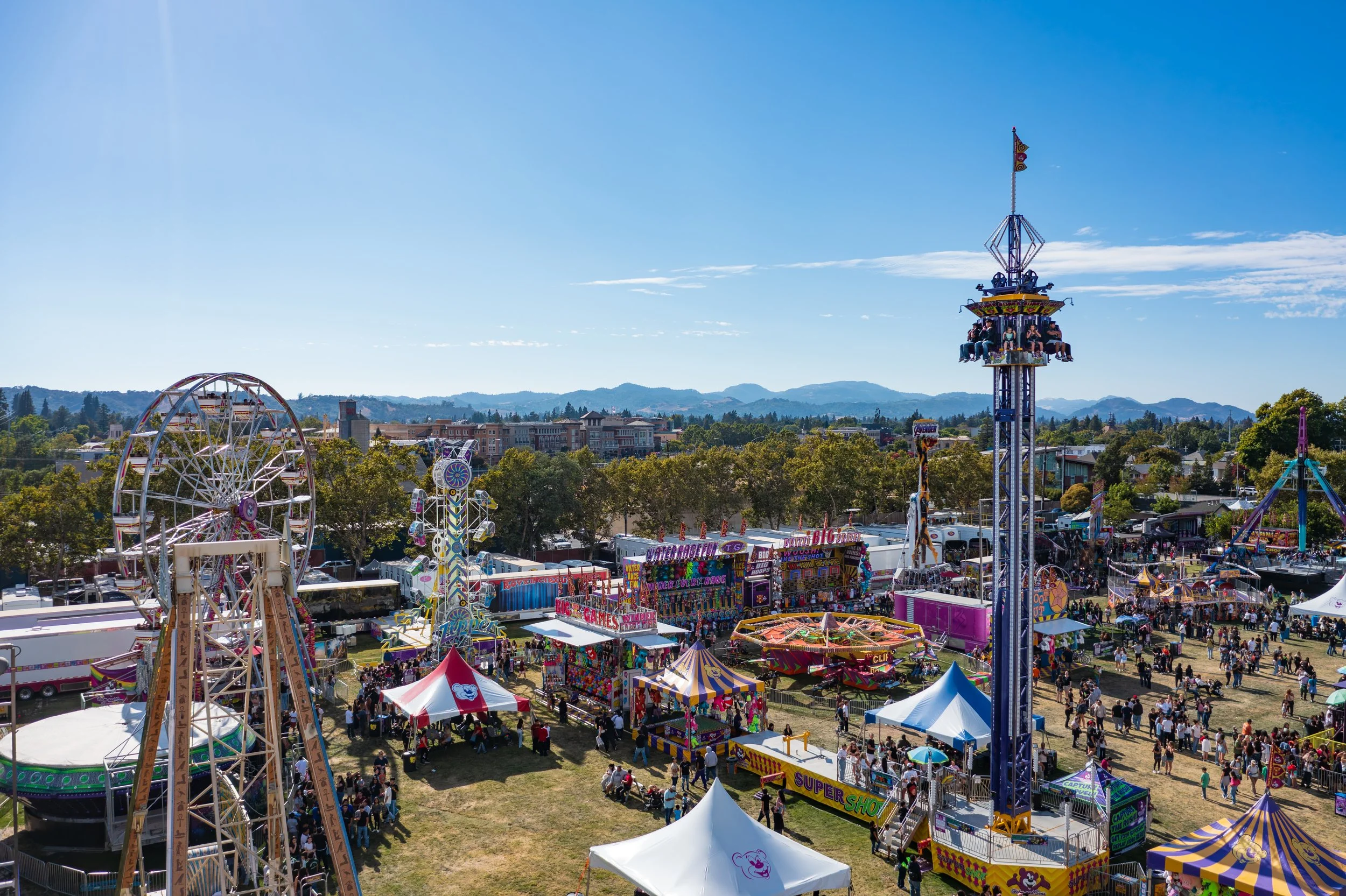 A wide view of a carnival with rides, game booths, and tents, with a mountain range in the background under a clear blue sky.