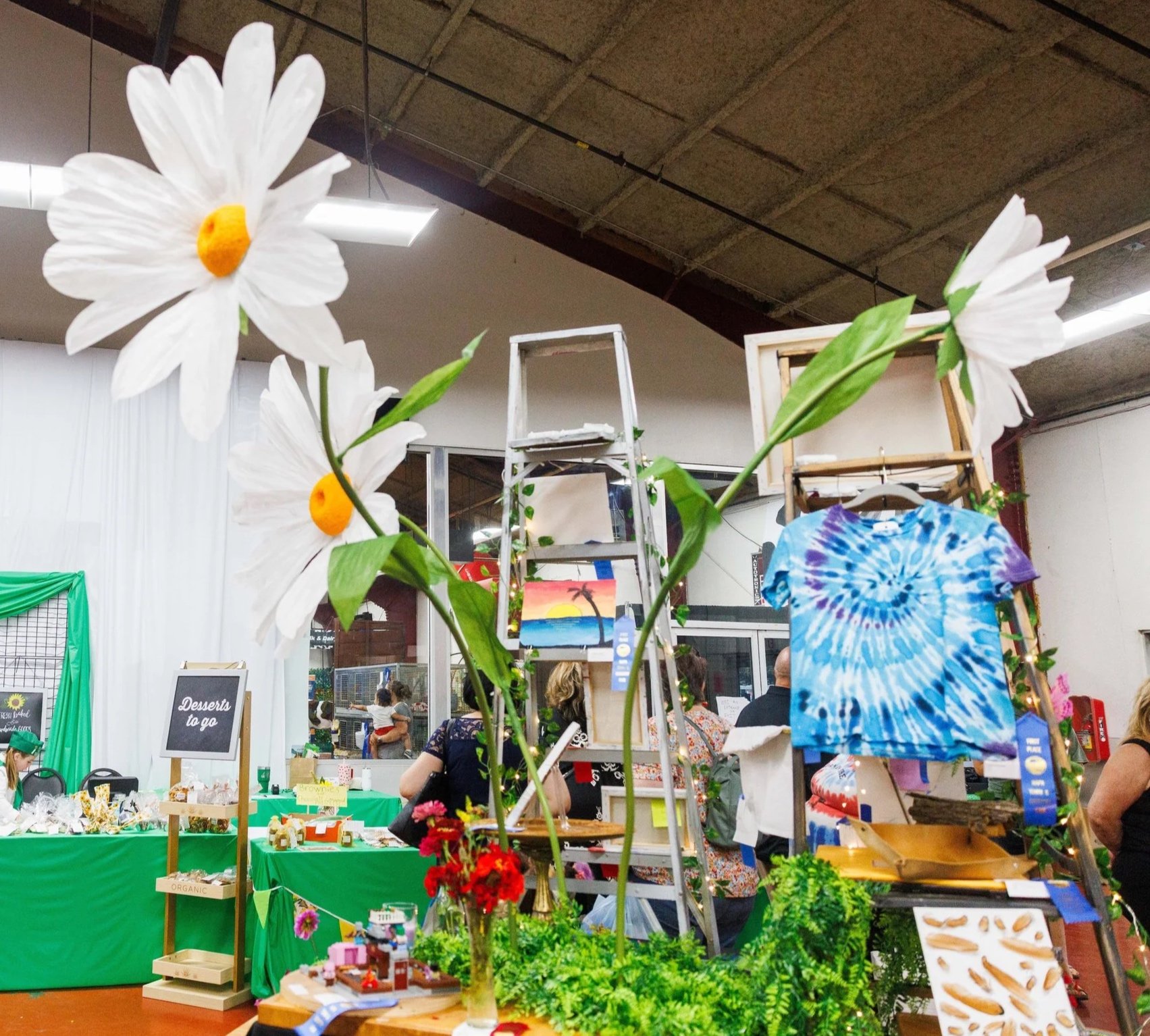 Indoor craft fair or market with various booths and displays. In the foreground, a large artificial white flower with a yellow center and green leaves. Background shows people shopping, colorful artwork, and handmade items on tables with green tablecloths. Industrial ceiling with exposed beams and lighting.
