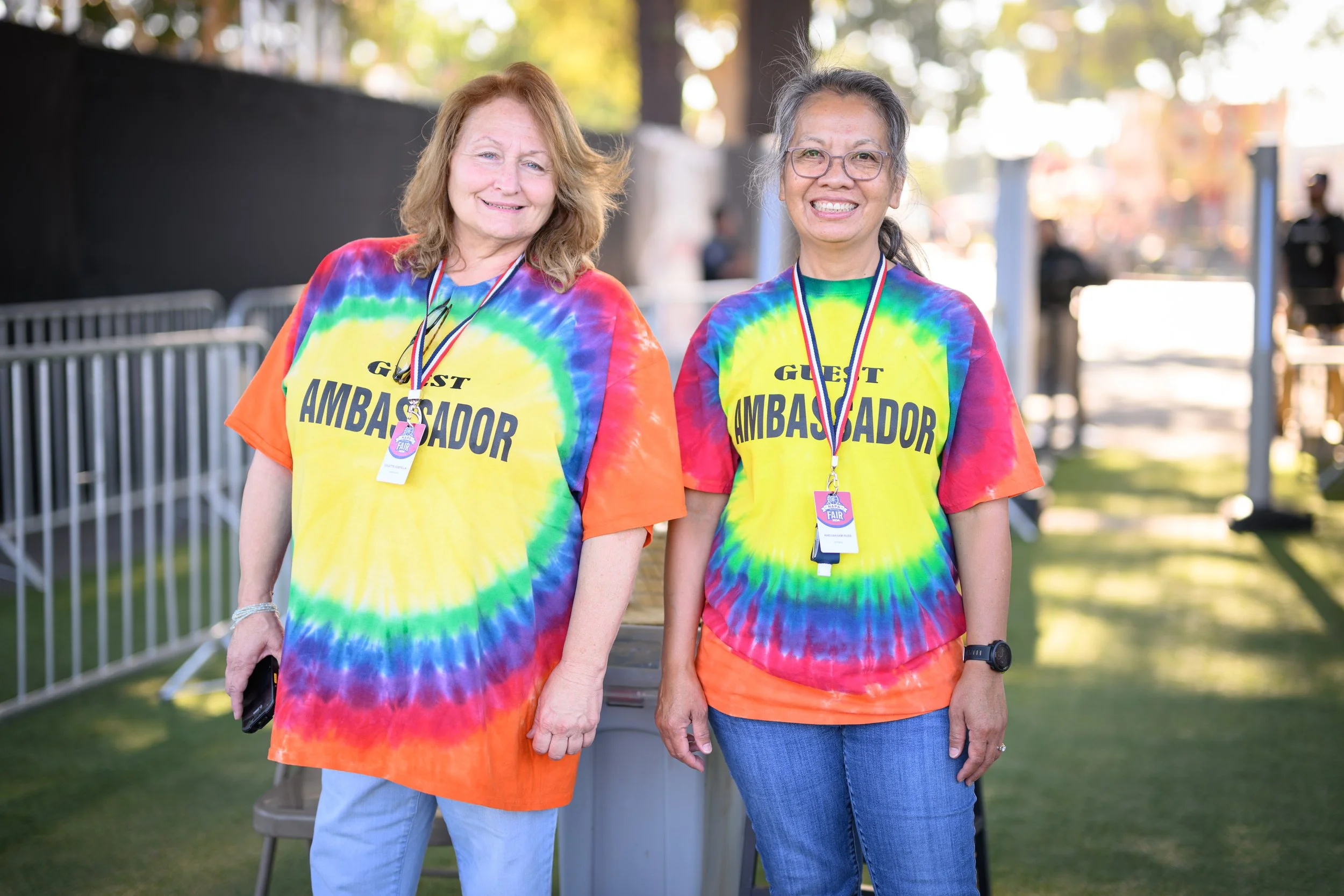 Two women walking at an outdoor event, wearing rainbow tie-dye T-shirts with 'Guest Ambassador' written on them, and medals around their necks, smiling at the camera.
