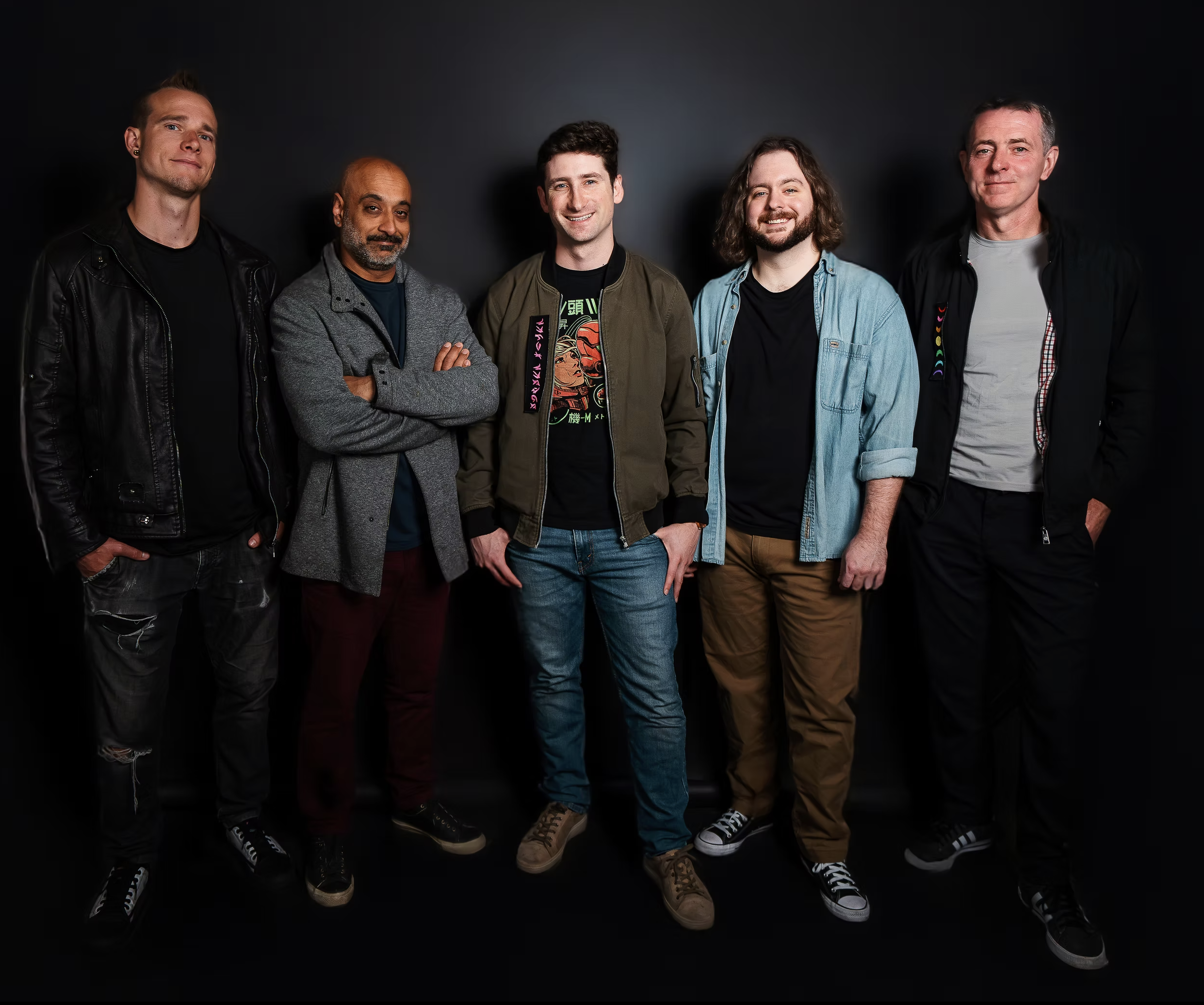 Group of six men standing against a dark background, smiling and posing for the photo. They are dressed casually, some with jackets and sneakers.