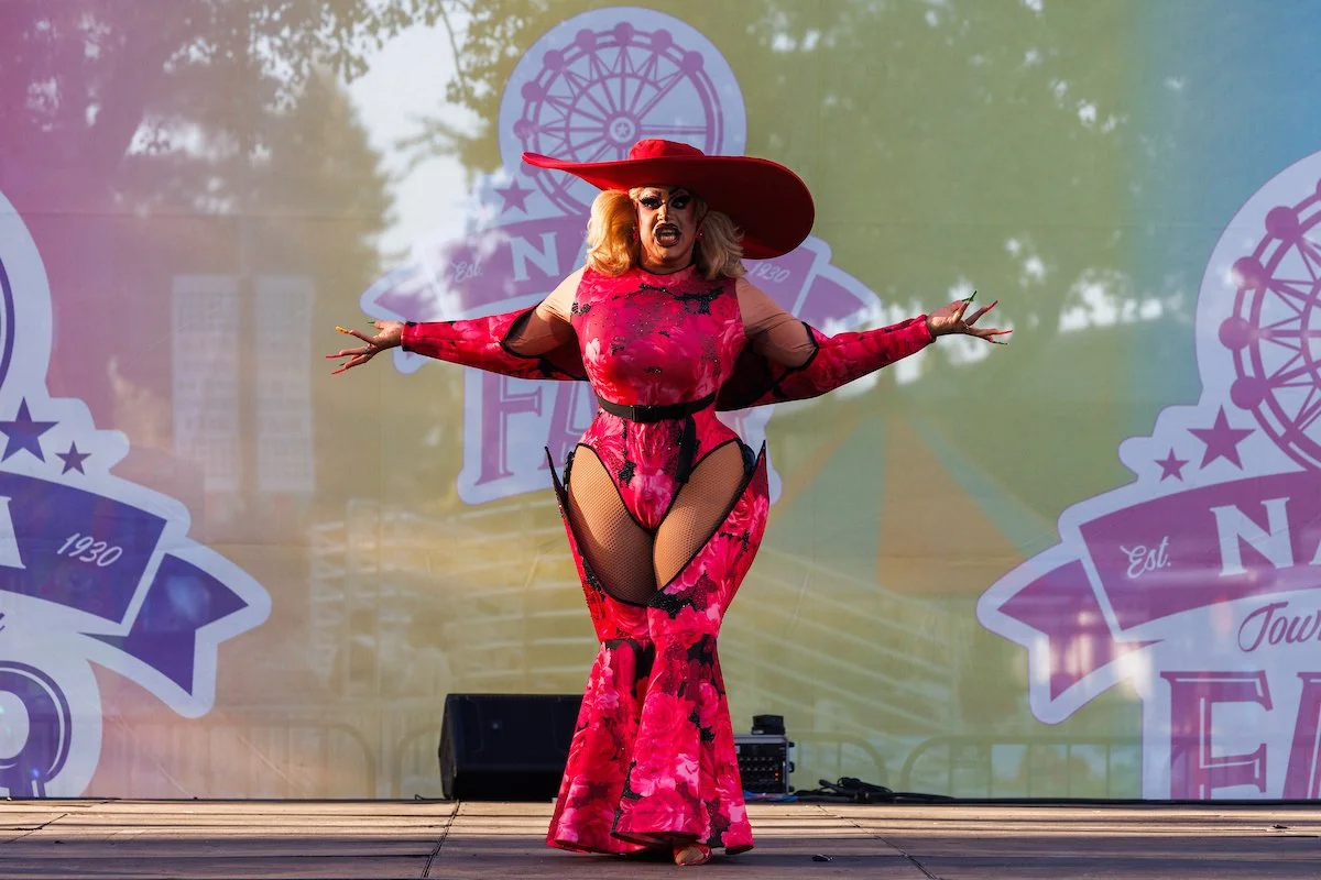 A drag performer dressed in a vibrant pink and black floral costume with sheer mesh and fishnet details, wearing a large red wide-brimmed hat, performs on an outdoor stage with a colorful background featuring carnival-type logos and trees.