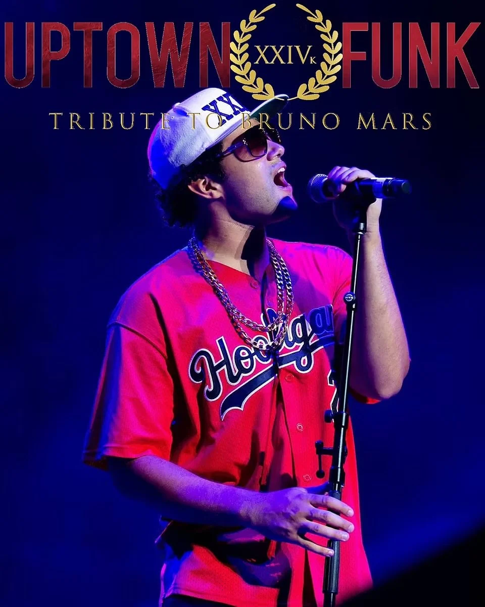 A man wearing a red sports jersey with 'Hoodie' across the front, sunglasses, a white cap, and gold chains, singing into a microphone on stage under blue and purple lights in tribute to Bruno Mars.