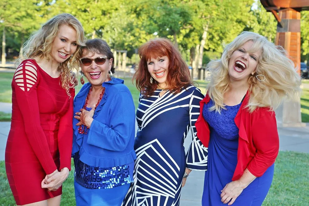 Four women smiling and laughing outdoors in a park with trees, dressed in vibrant dresses and jackets.