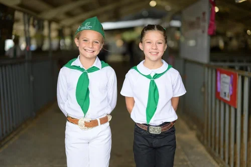 Two young girls dressed in Cub Scout uniforms standing outdoors, smiling at the camera.