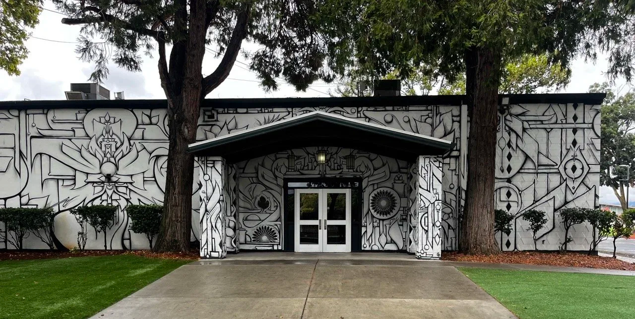 Building with black and white geometric mural art, surrounded by trees and bushes, with a concrete walkway leading to glass double doors.