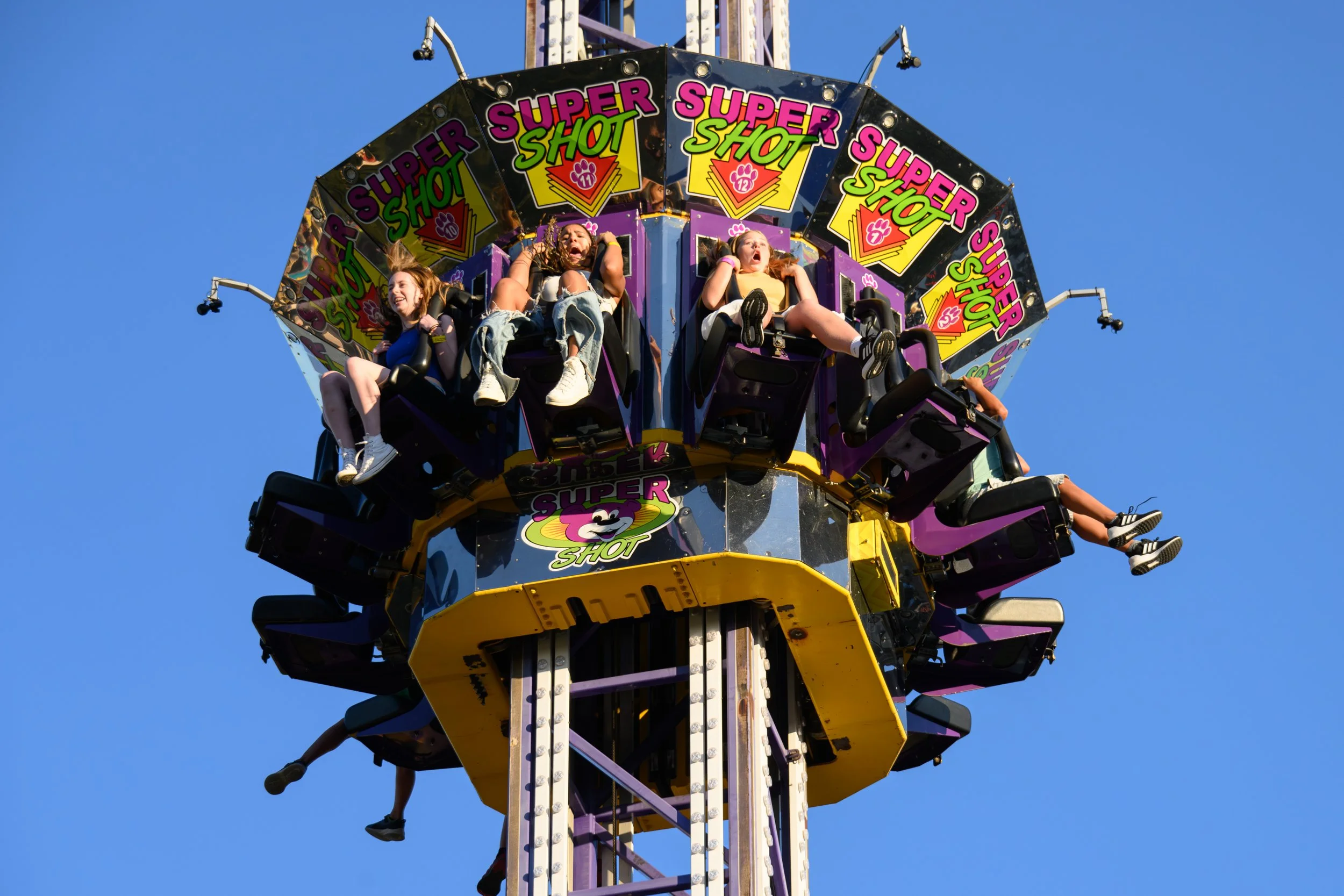Three young girls on a colorful spinning amusement park ride named 'Super Shot,' seen against a clear blue sky.