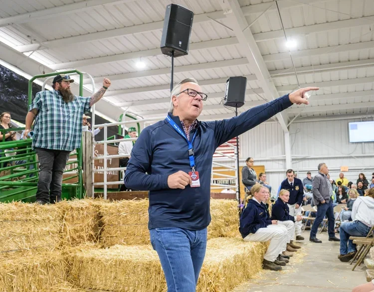 A man wearing glasses and a navy jacket is pointing to the right while speaking aloud at a farming event inside a barn, with hay bales and other people, including children, in the background.