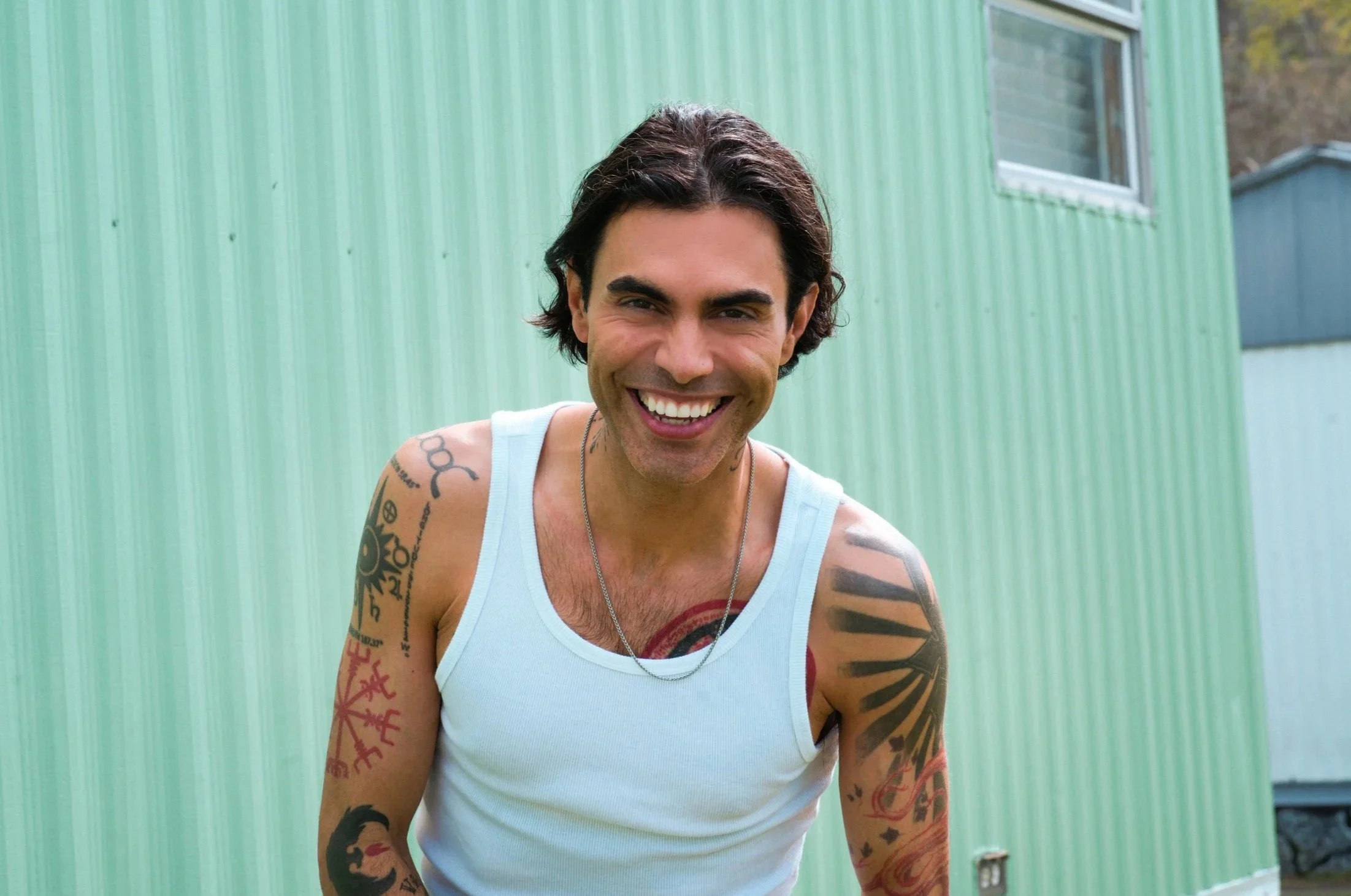 A man with dark, wavy hair and multiple tattoos on his arms, smiling outdoors in front of a green building with a window.