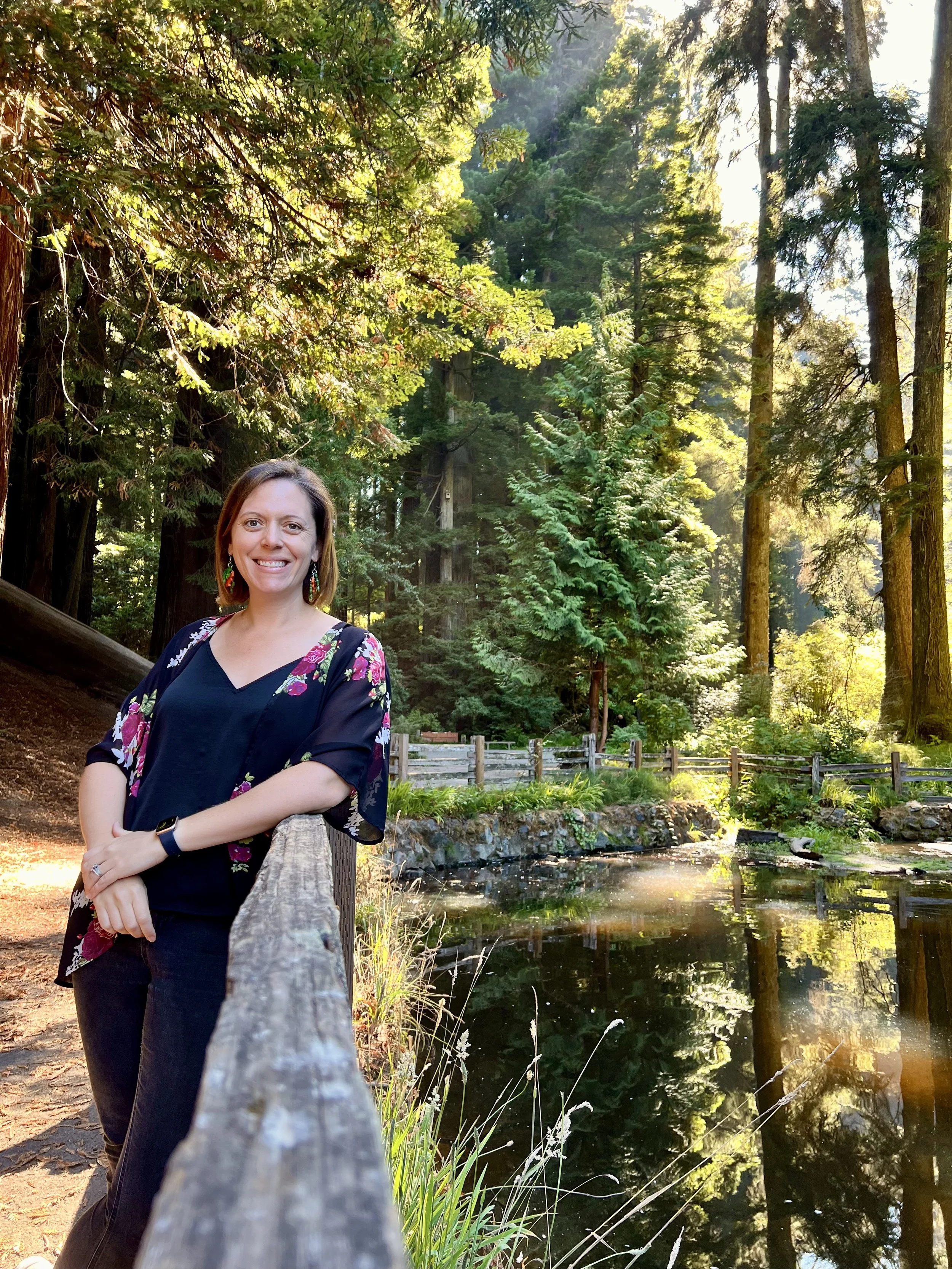 Woman smiling and standing by a wooden fence along a pond in a lush, green forest with tall trees and sunlight filtering through the foliage.