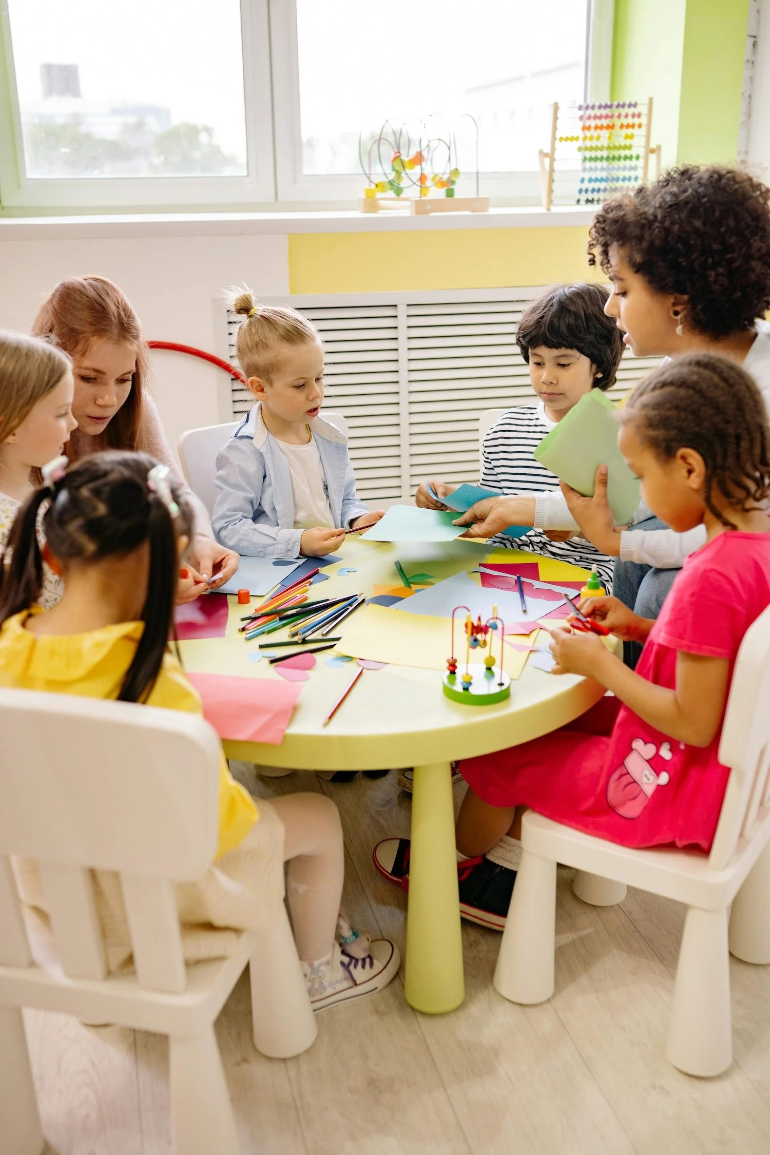 Group of young children and an adult woman at a round table creating arts and crafts with colored paper, scissors, and markers in a brightly lit room.