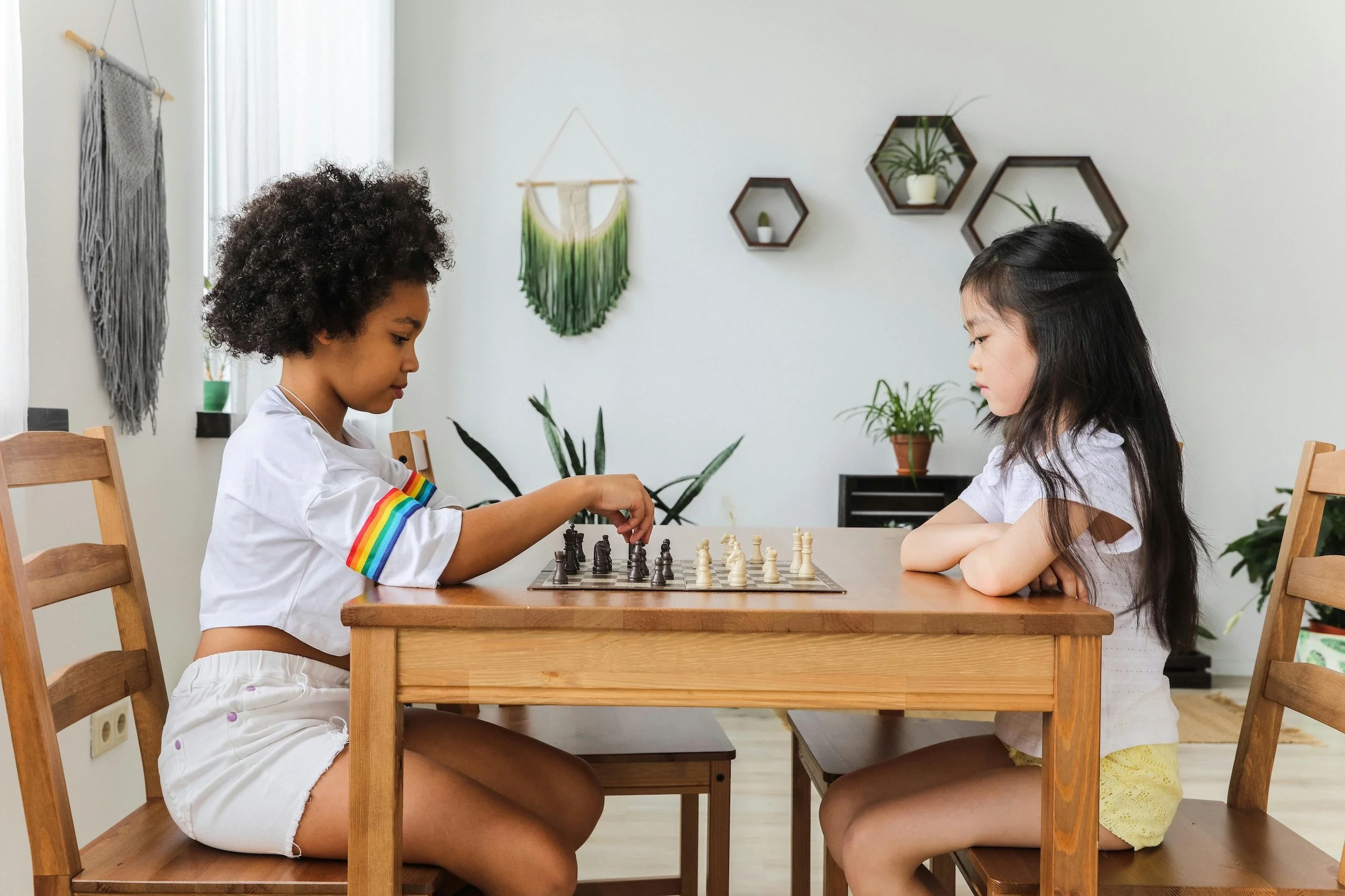 Two young girls playing chess at a wooden table in a bright room with plants and wall decorations.