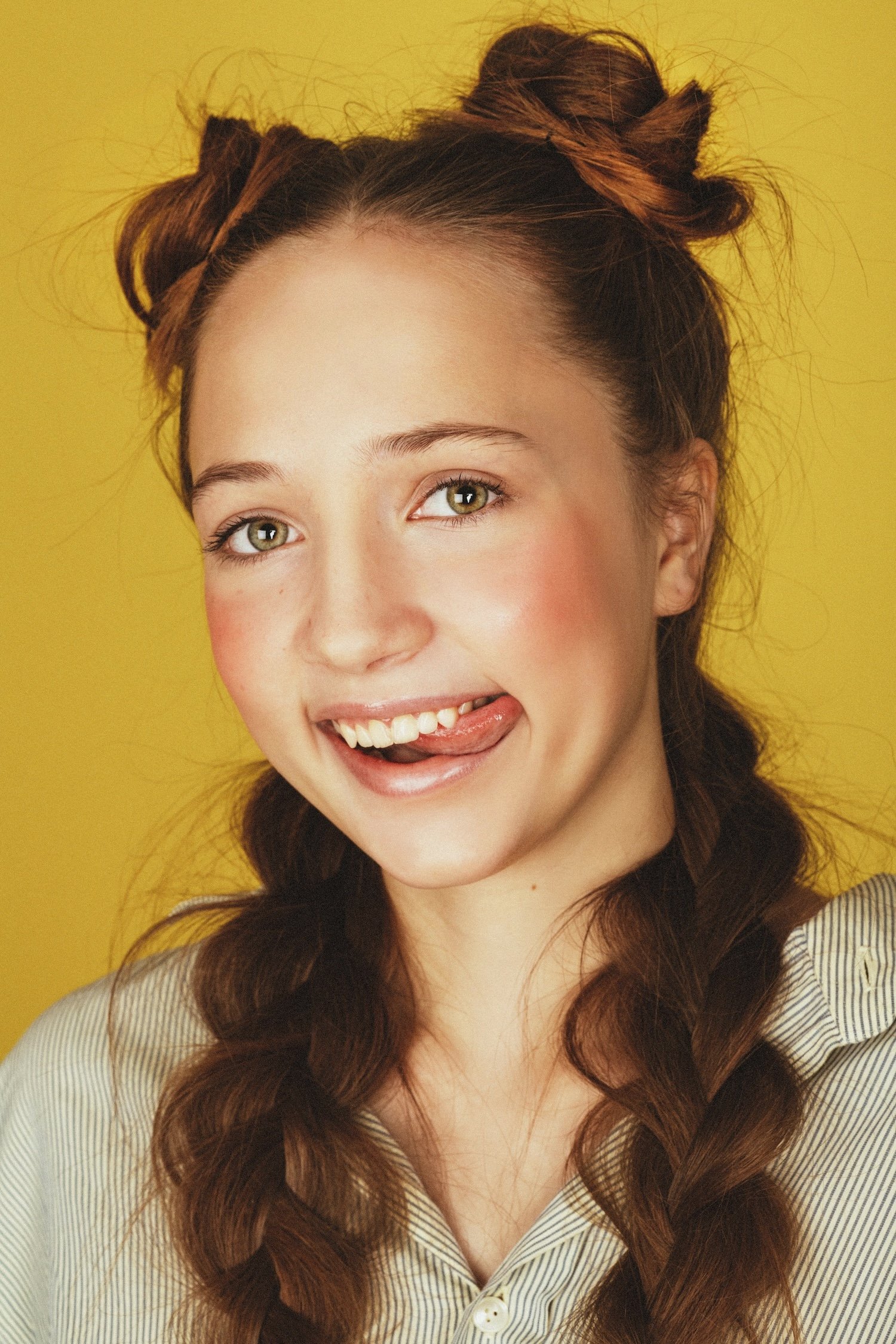A young woman with light skin and green eyes making a playful face, sticking out her tongue and tilting her head, against a yellow background.