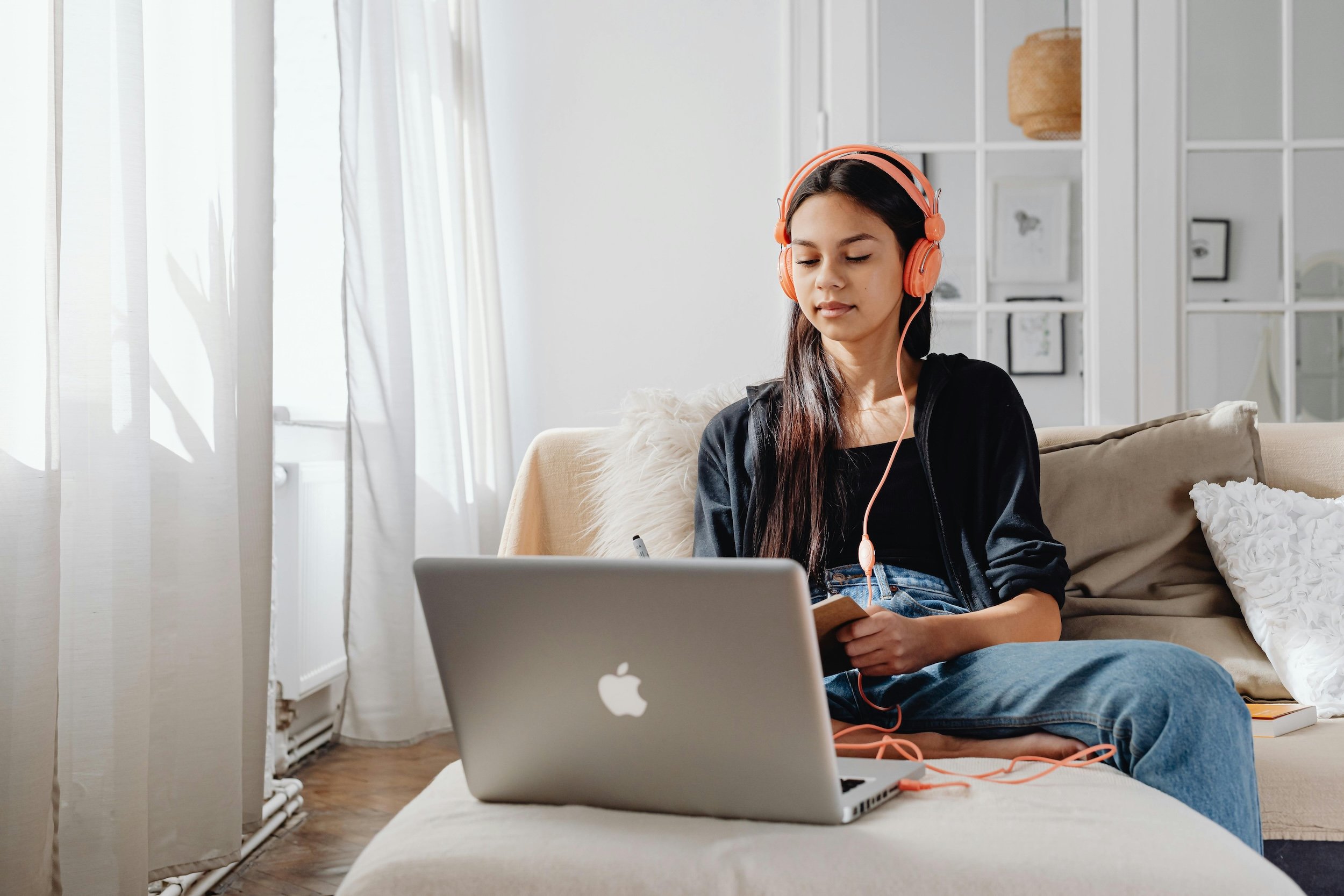 A young woman sitting on a beige sofa, wearing pink over-ear headphones, working on a laptop, and holding a notebook in a bright, modern room.
