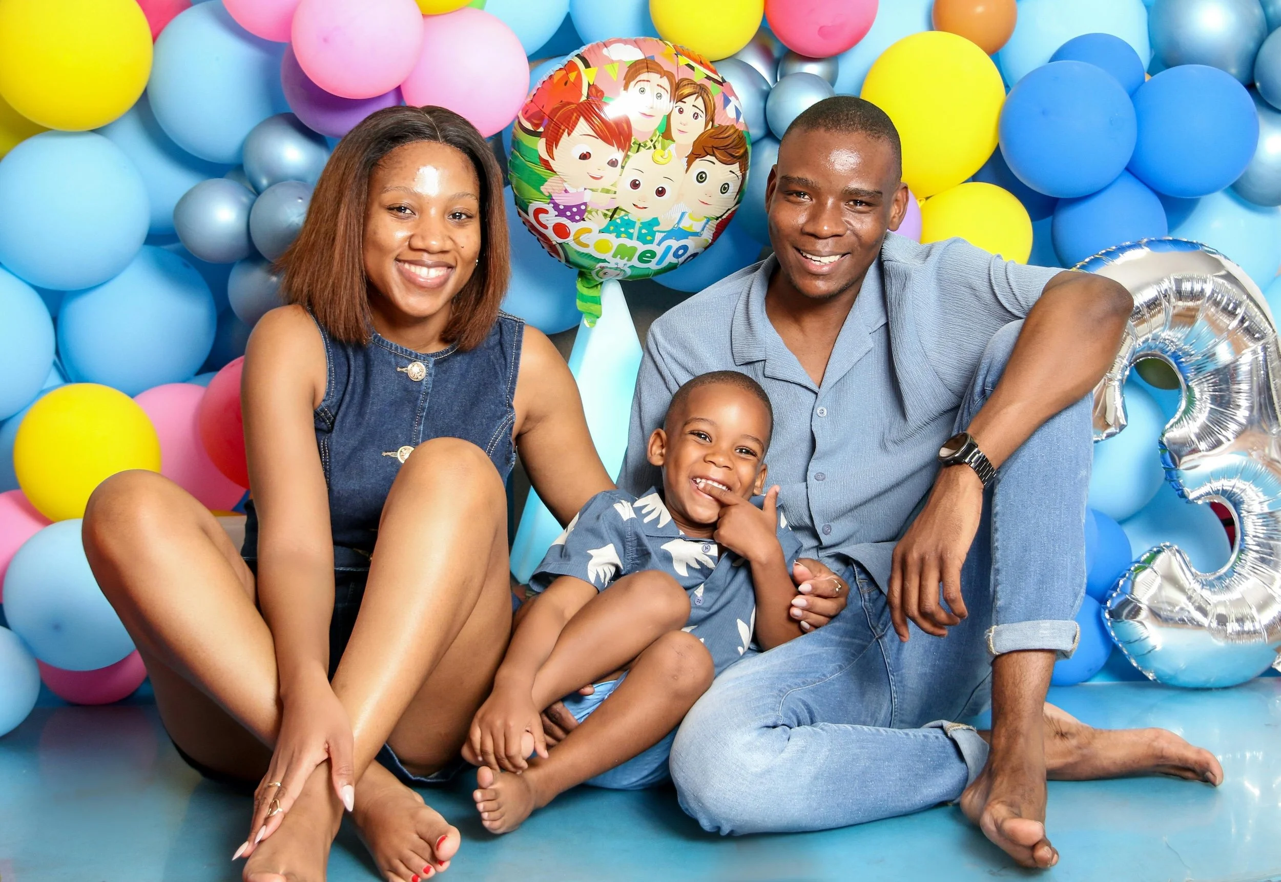 A smiling family of three sitting on blue floor in front of a colorful balloon backdrop with a 'Paw Patrol' themed balloon. The family includes a woman with shoulder-length hair in a blue denim dress, a young boy in a patterned shirt, and a man in a light blue shirt and jeans. They appear happy and relaxed.