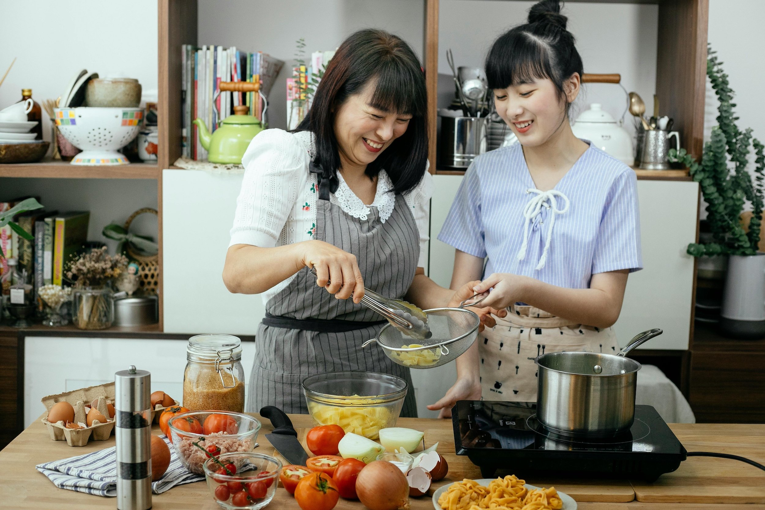 A woman and a girl cooking together in a kitchen, with various vegetables and cooking ingredients on the counter.