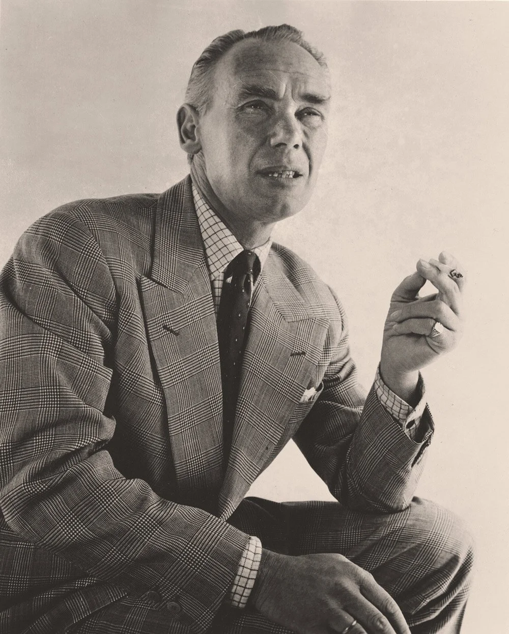 Black and white photograph of a man wearing a checked suit with a matching tie. He is sitting and holding a cigarette in his right hand, with a serious expression.