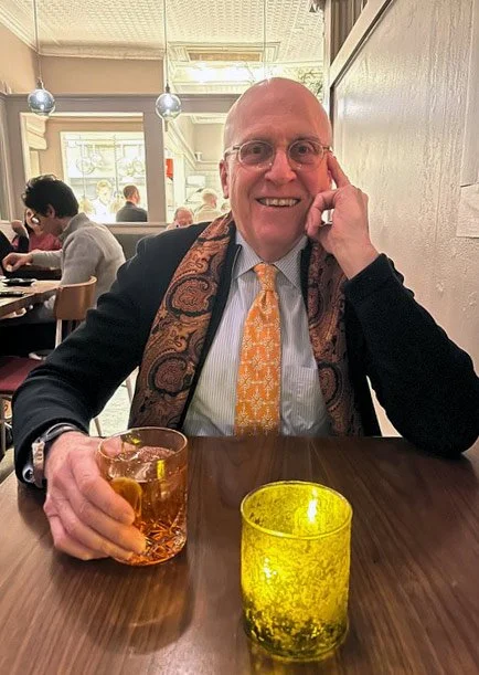 A smiling man with glasses and a bald head sitting at a restaurant table, holding a glass of a brown drink, with lit candles on the table, and other patrons in the background.