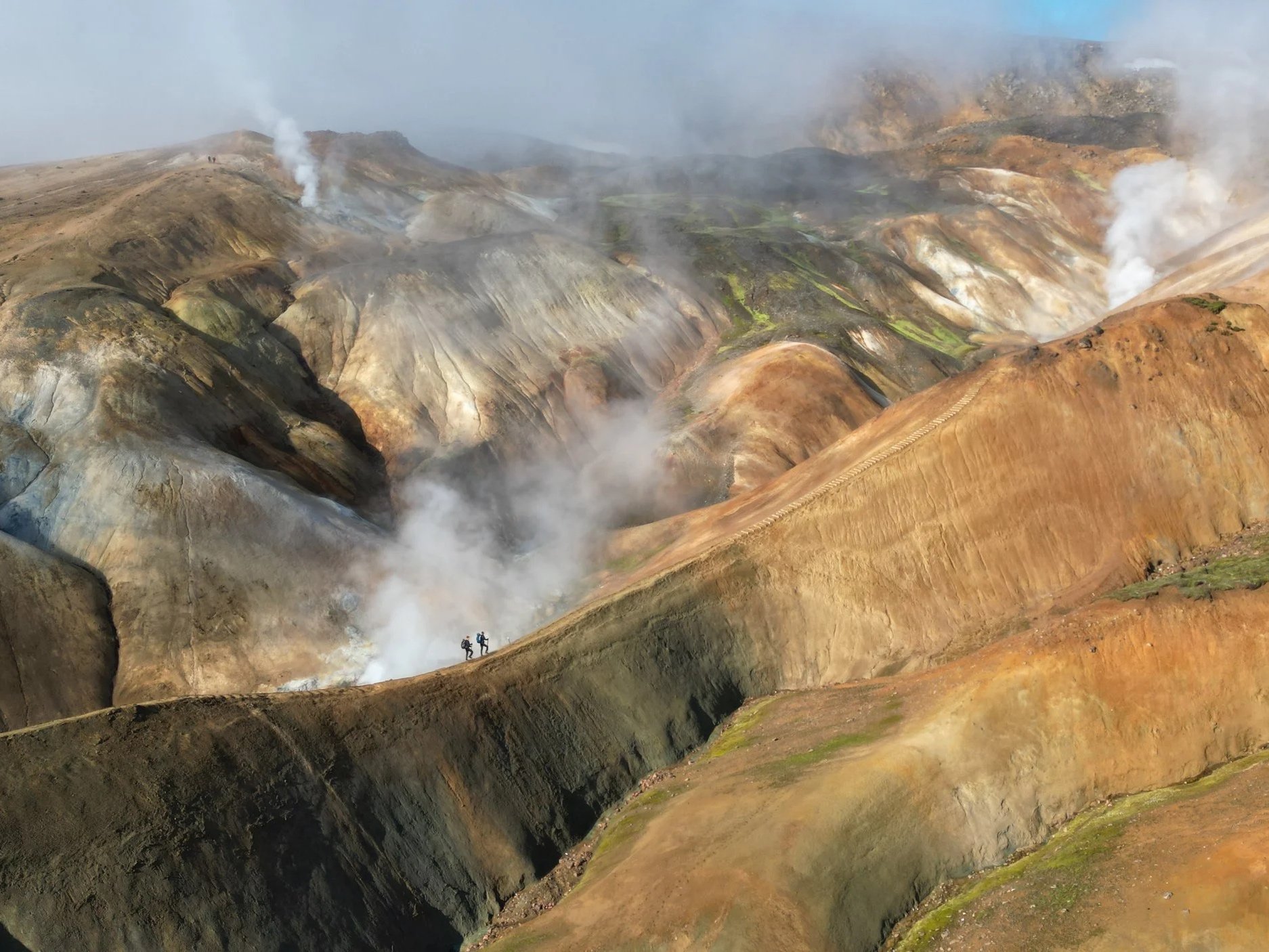 Aerial view of hikers walking along a volcanic ridge, showing scale within an active geothermal landscape.