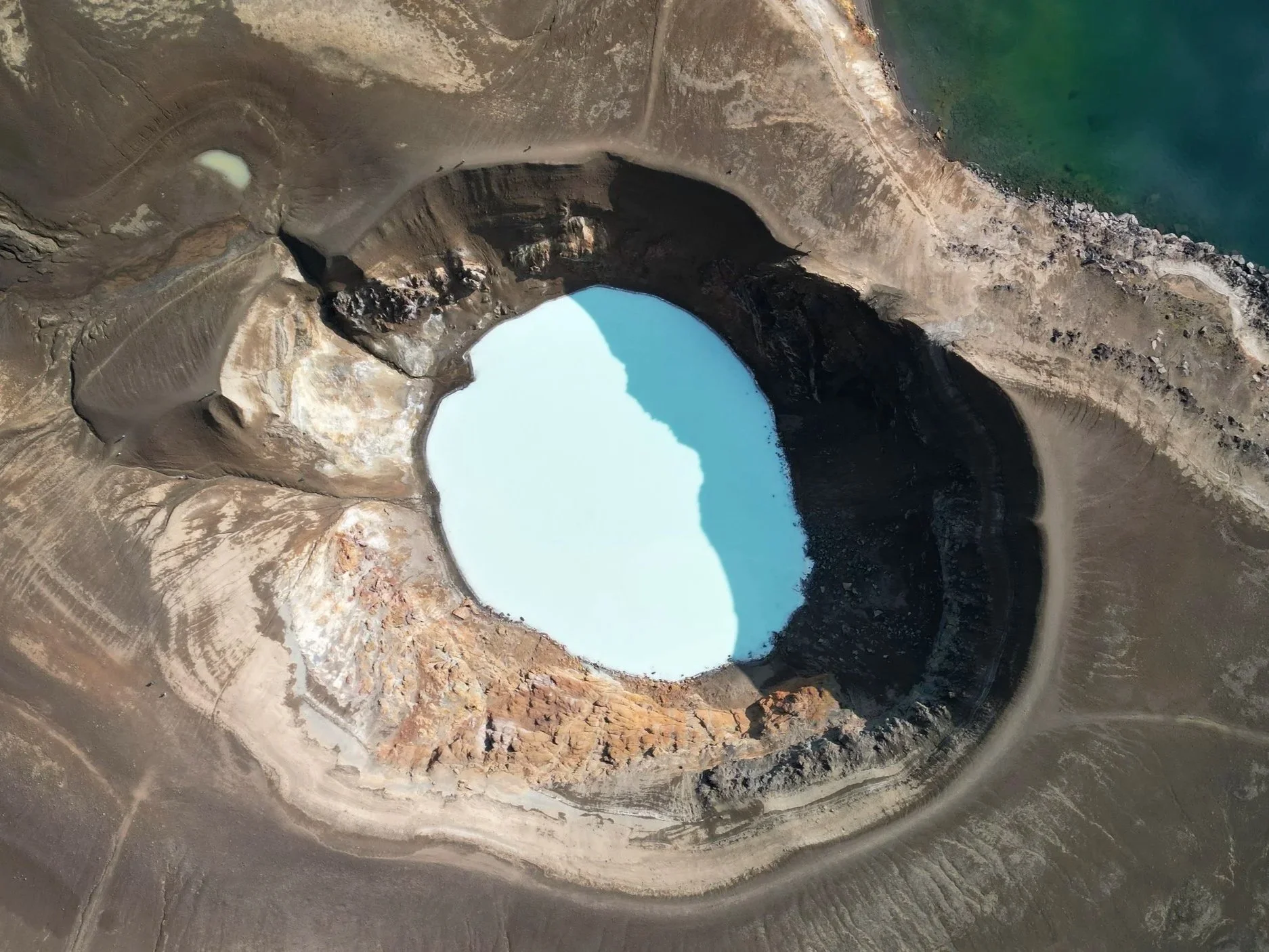 Aerial view of a volcanic crater with pale blue water, showing circular landform and surrounding barren terrain.