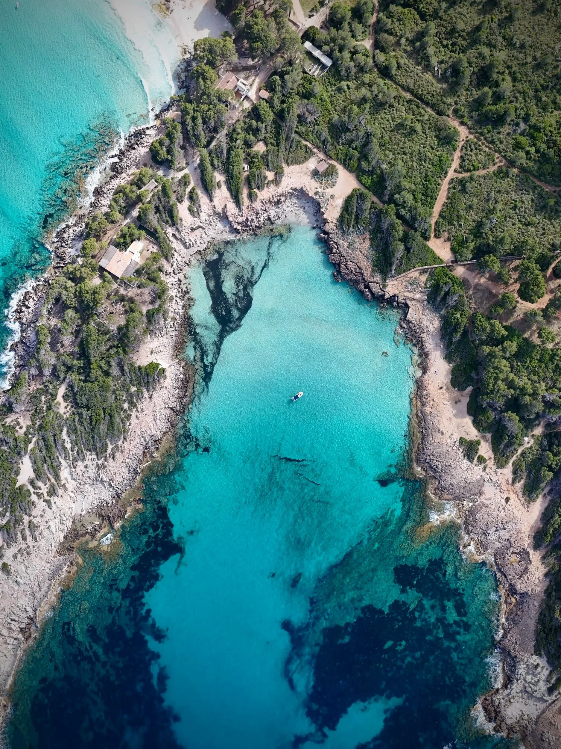Aerial view of a turquoise coastal cove surrounded by rocky shoreline and forested hills, showing clear water and coastal formations in Mallorca, Spain.