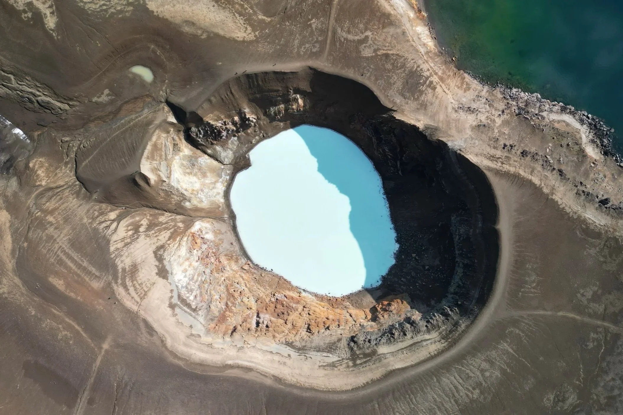Aerial view of a water reservoir or lake with a partially dried or ice-covered surface, surrounded by rugged, barren land and a body of water nearby.