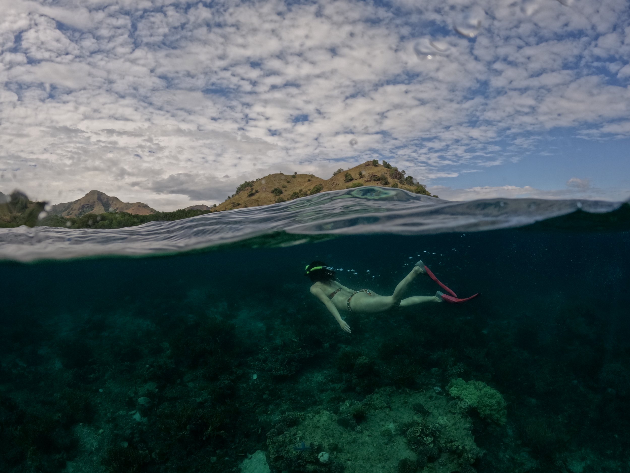 Half-underwater photograph of a swimmer above coral reef with island landscape in Komodo National Park.