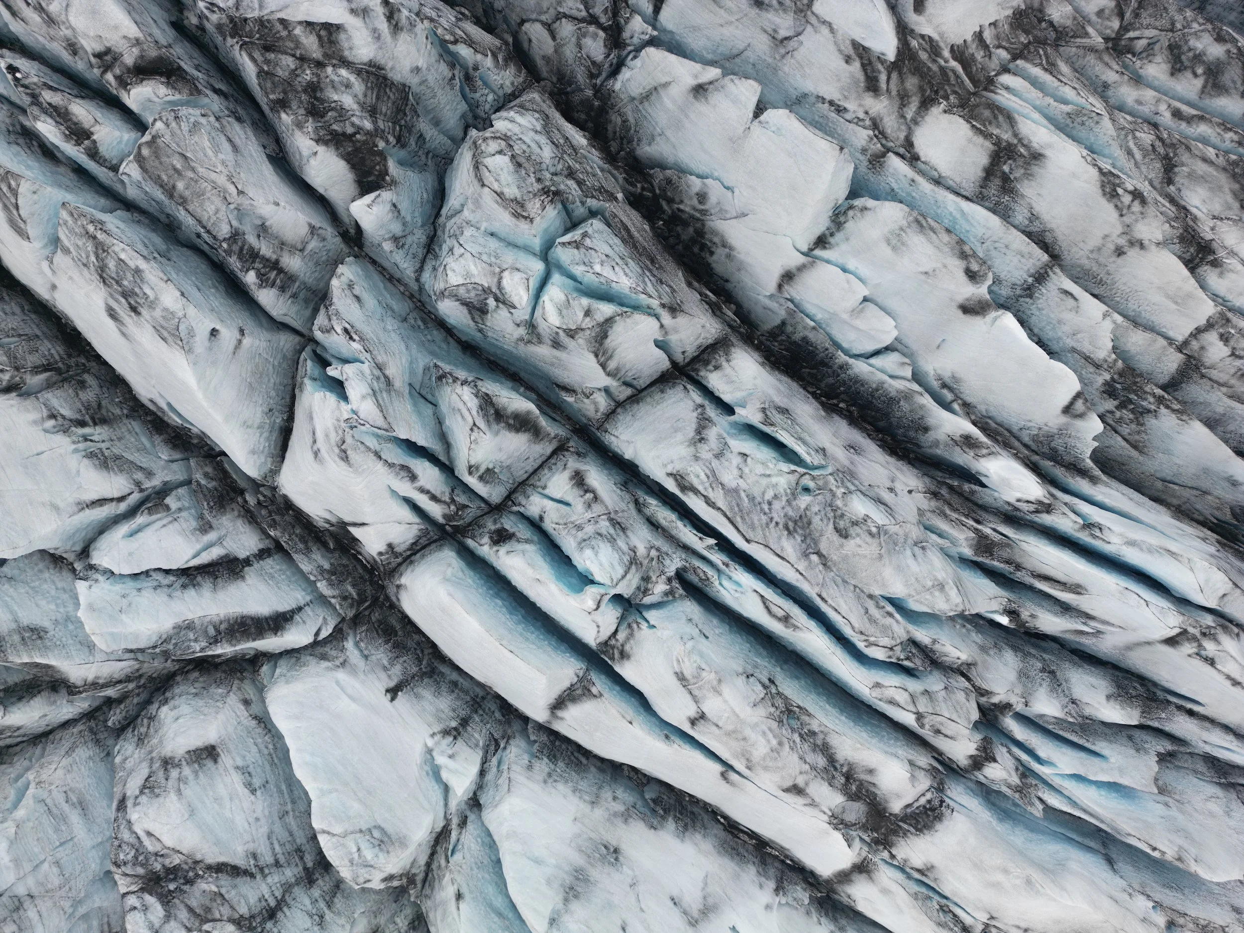 Aerial view of glacial ice formations with crevasses and flowing meltwater, showing abstract patterns in white and blue tones.